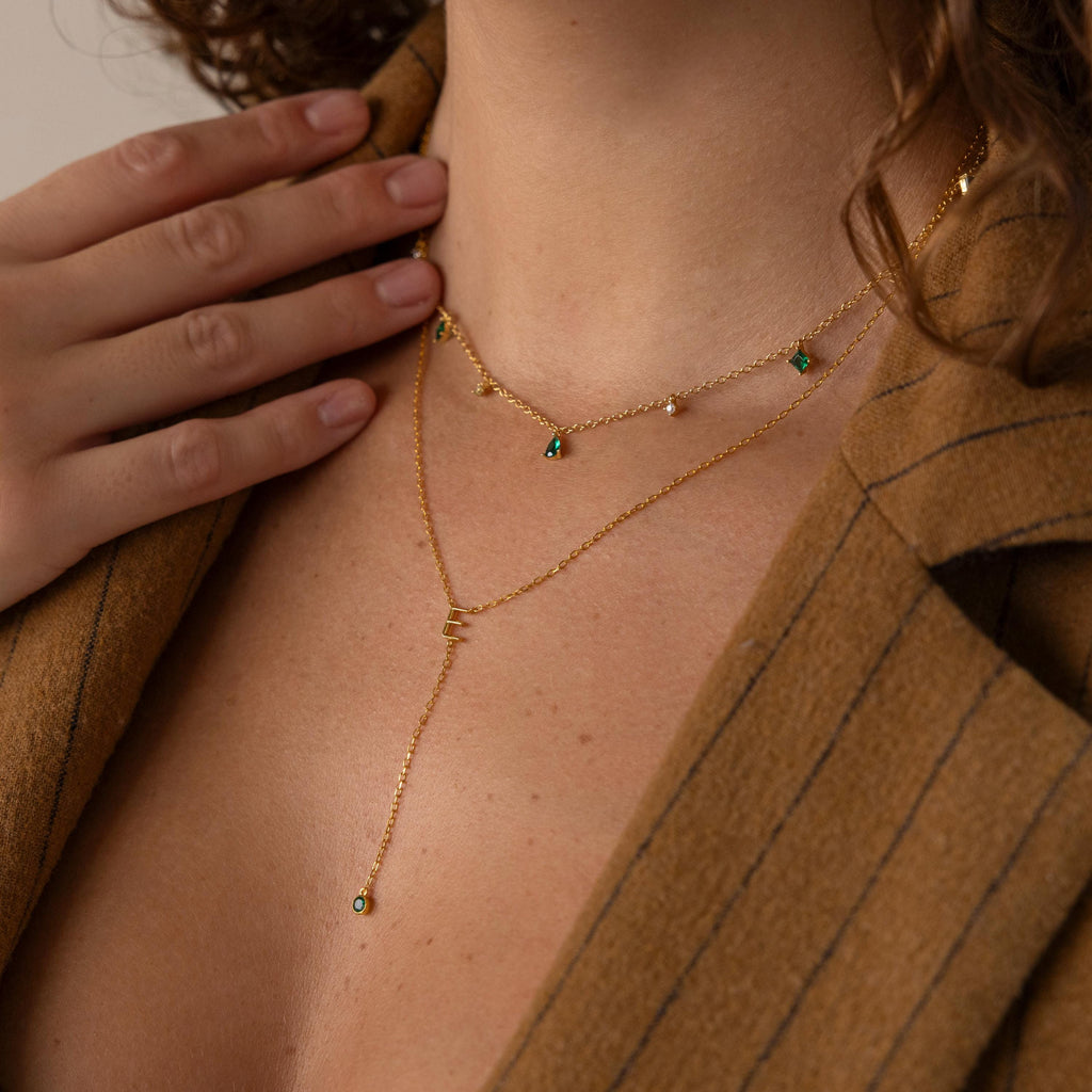 A woman in a tan blazer with a deep neckline wears layered gold necklaces, including the Initial Birthstone Lariat Necklace featuring green stones.