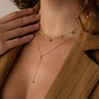 A woman in a tan blazer with a deep neckline wears layered gold necklaces, including the Initial Birthstone Lariat Necklace featuring green stones.