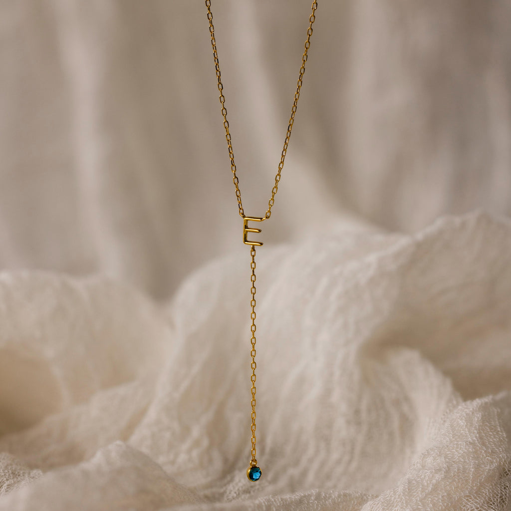 Initial Birthstone Lariat Necklace: Gold lariat necklace featuring an E initial pendant and a blue gemstone drop, shown against a soft cream fabric background.