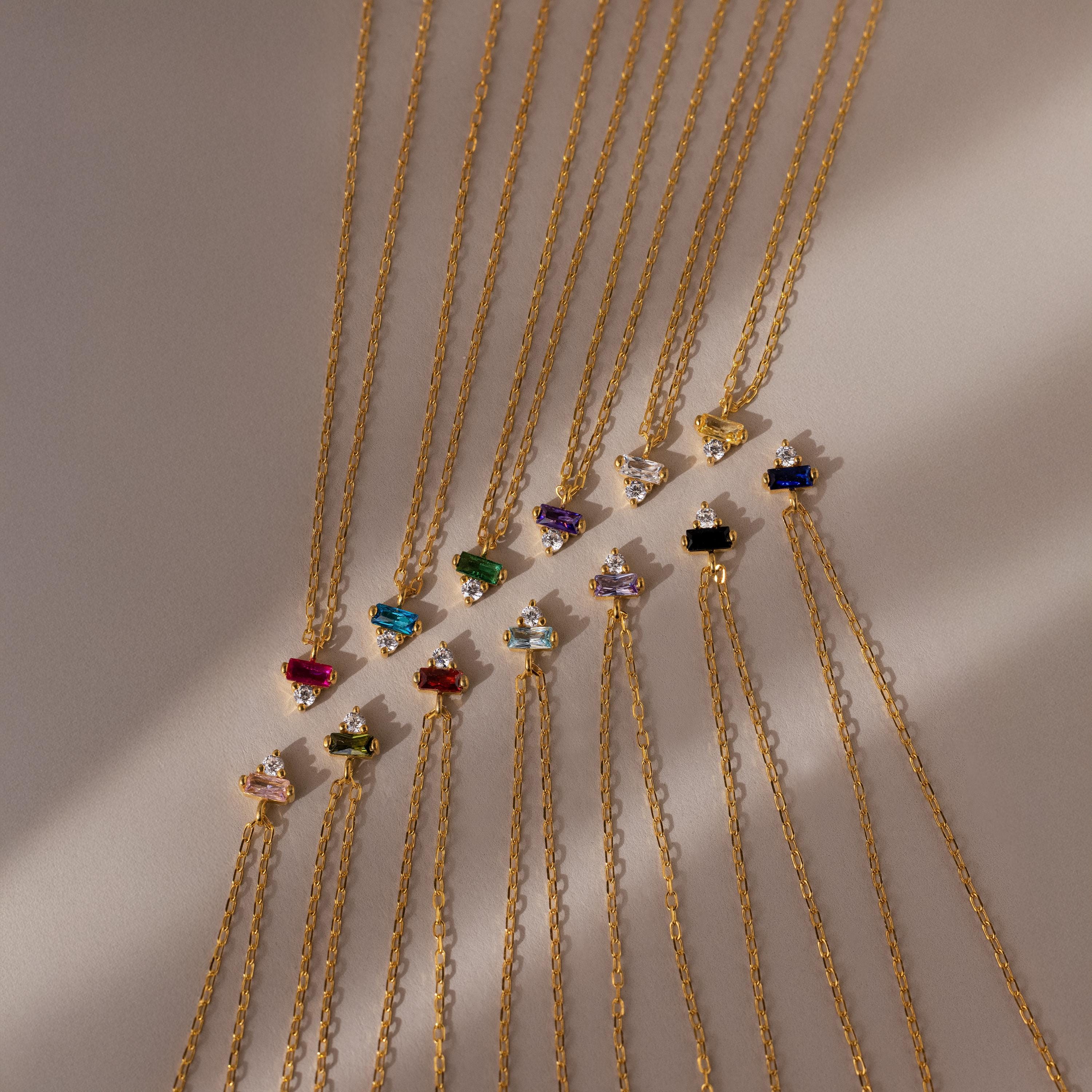 Baguette Birthstone Drop Necklaces, featuring colorful gemstone pendants, are arranged in neat diagonal rows on a light surface.
