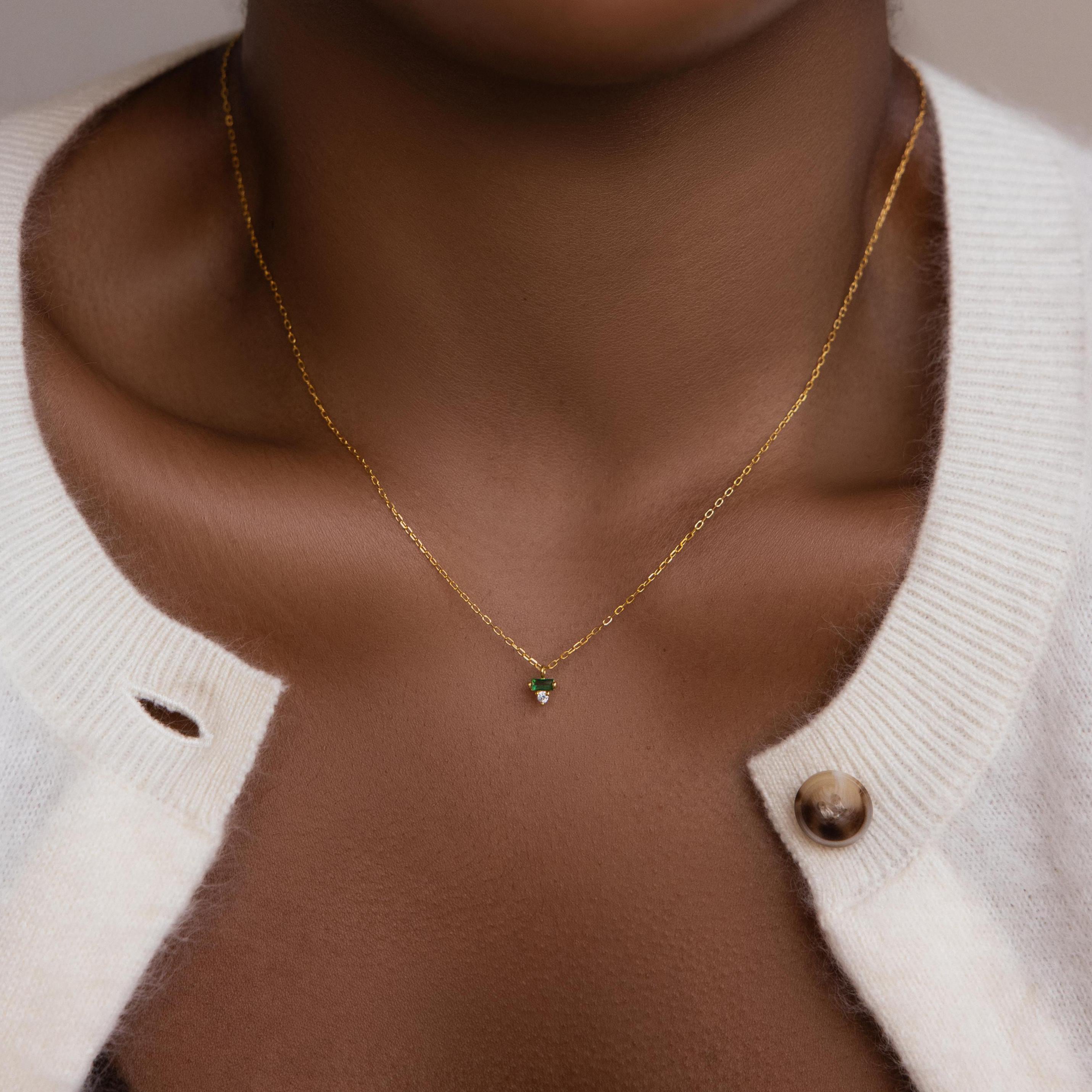 A close-up of a Baguette Birthstone Drop Necklace featuring a small green pendant, worn by someone in a white sweater.
