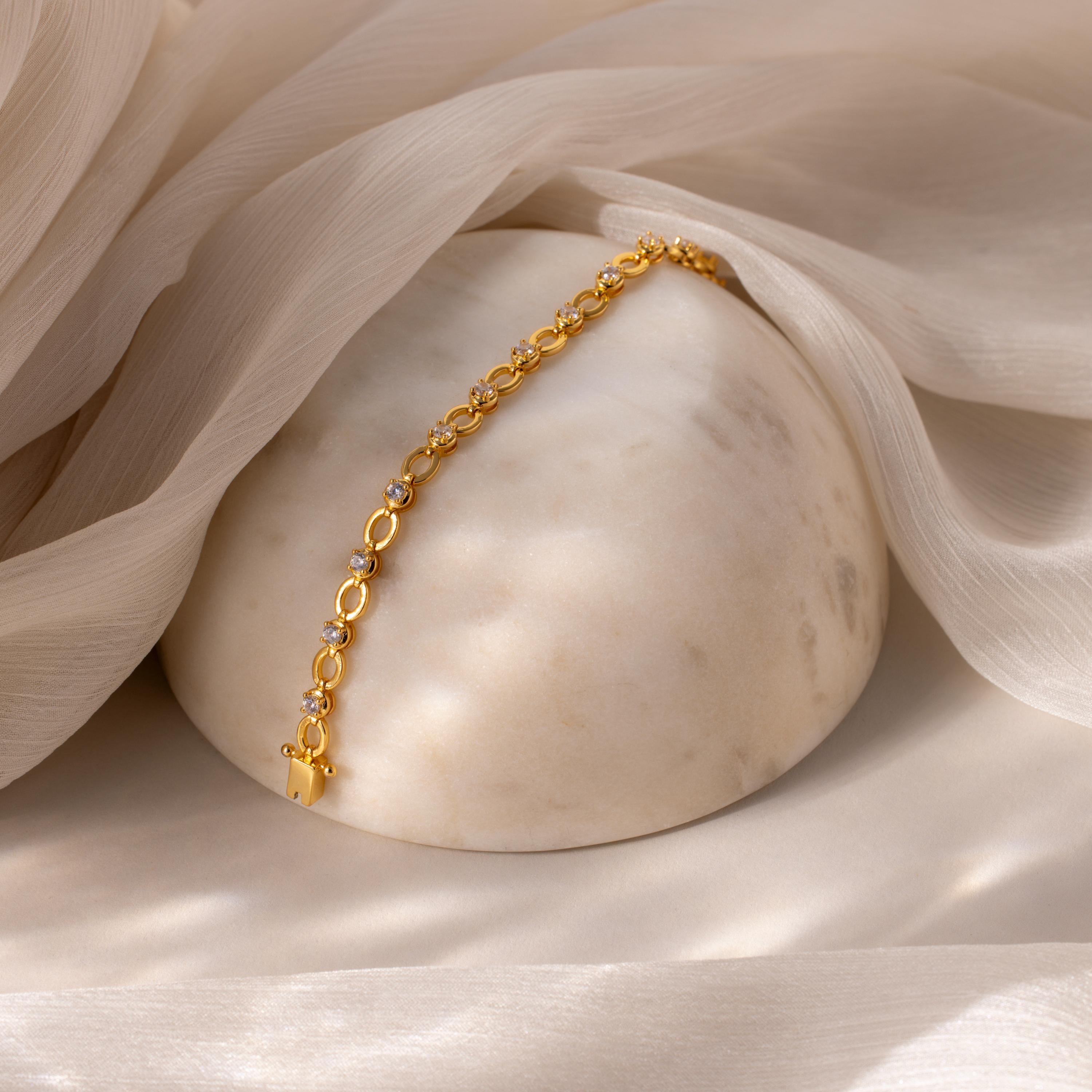 The Diamond Chain Tennis Bracelet, featuring gold links and round accents, is displayed on smooth marble and draped with sheer beige fabric.