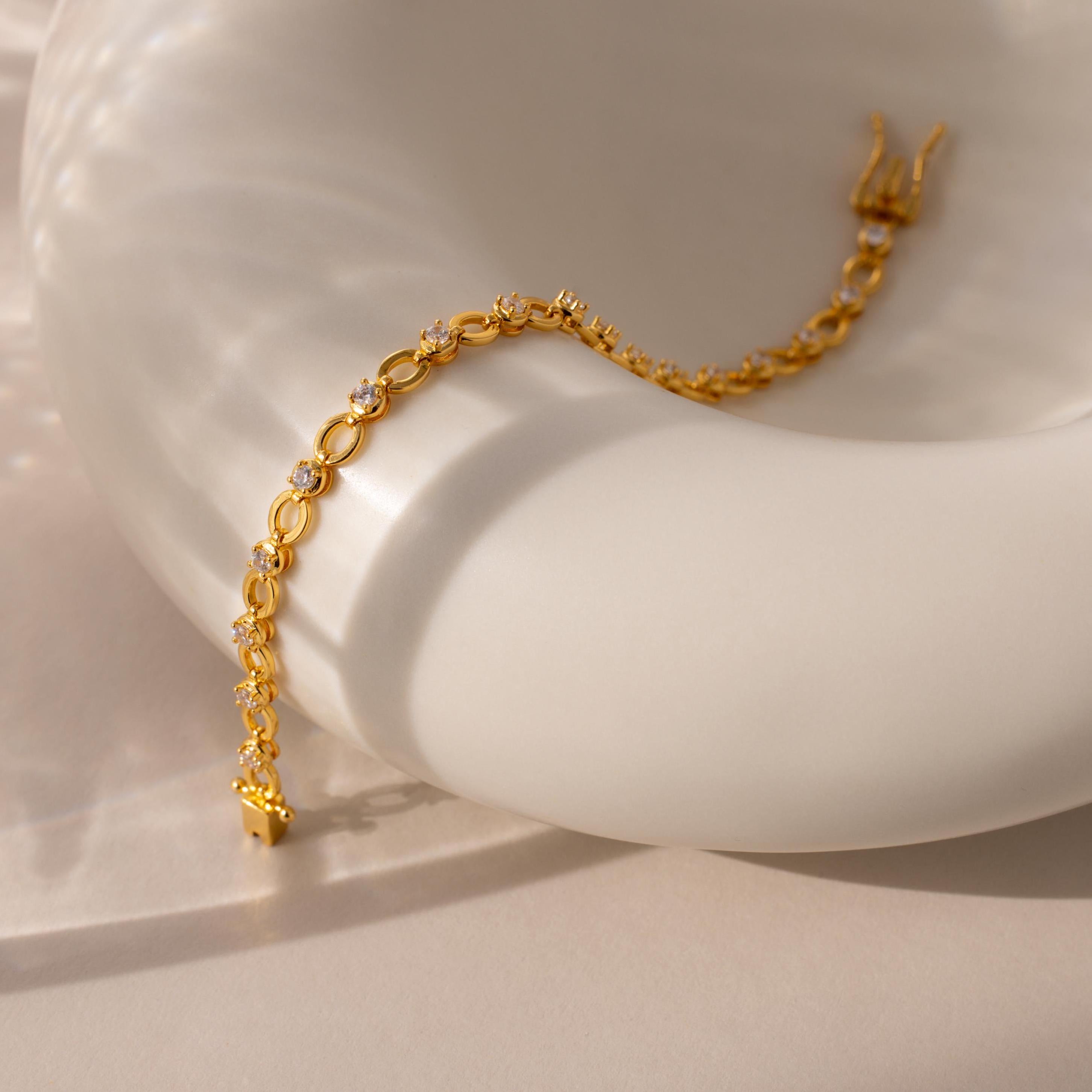 The Diamond Chain Tennis Bracelet, featuring gold links and clear diamonds, rests elegantly on a sleek white ceramic surface under gentle lighting.