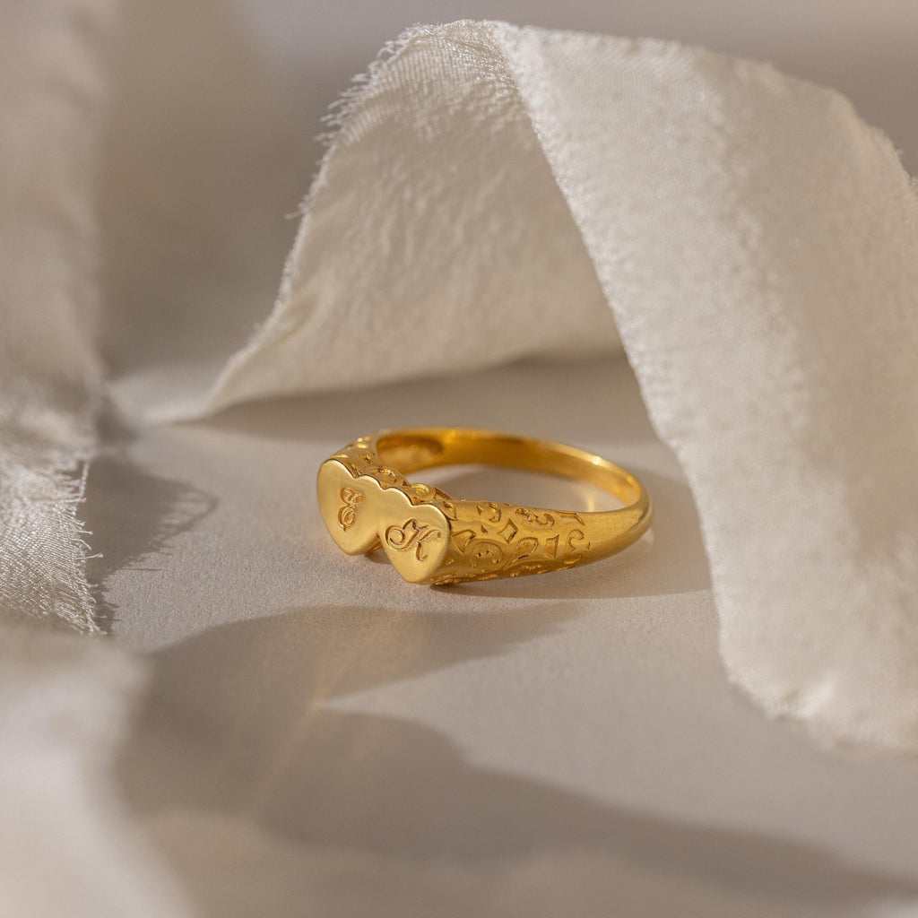 The Duo Initial Heart Ring features two heart shapes engraved with initials, beautifully displayed on soft, white fabric.