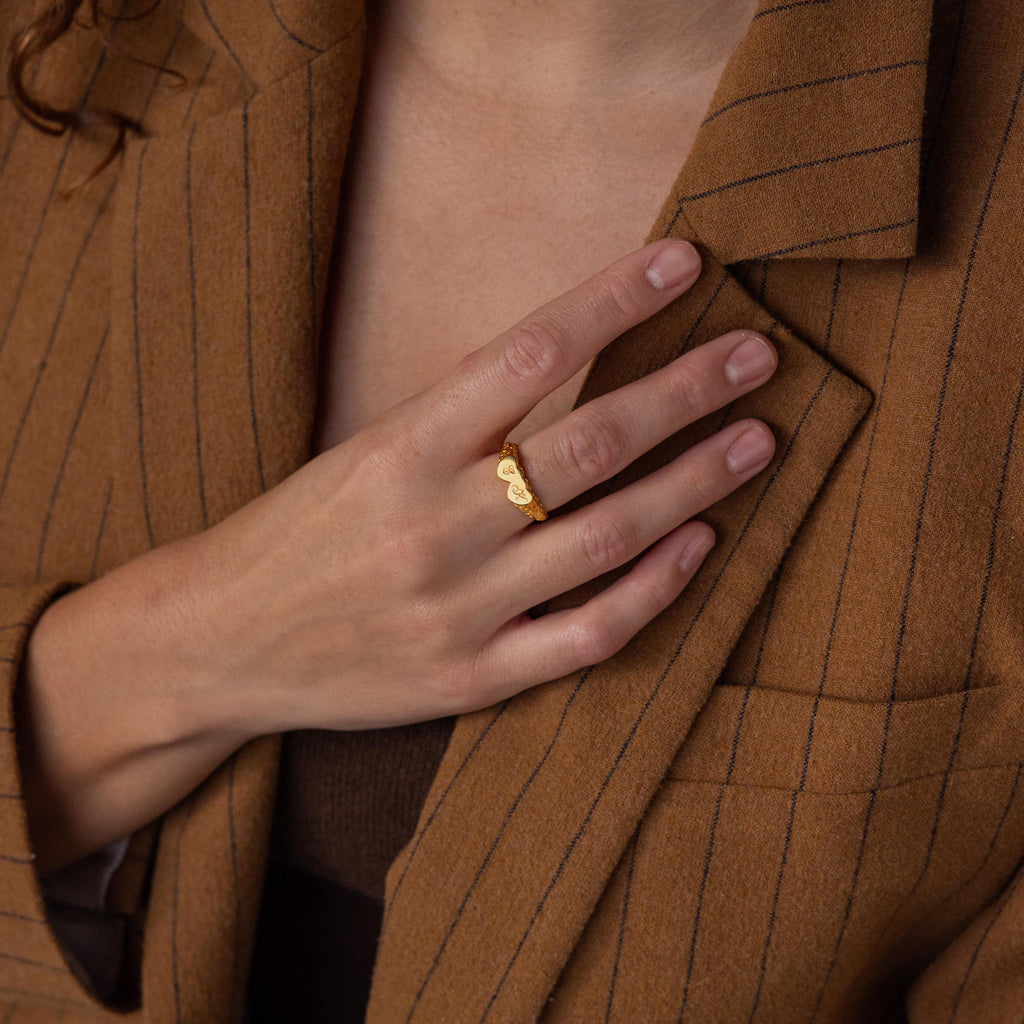 A hand with neatly manicured nails showcases the Duo Initial Heart Ring while resting on a brown pinstripe blazer.