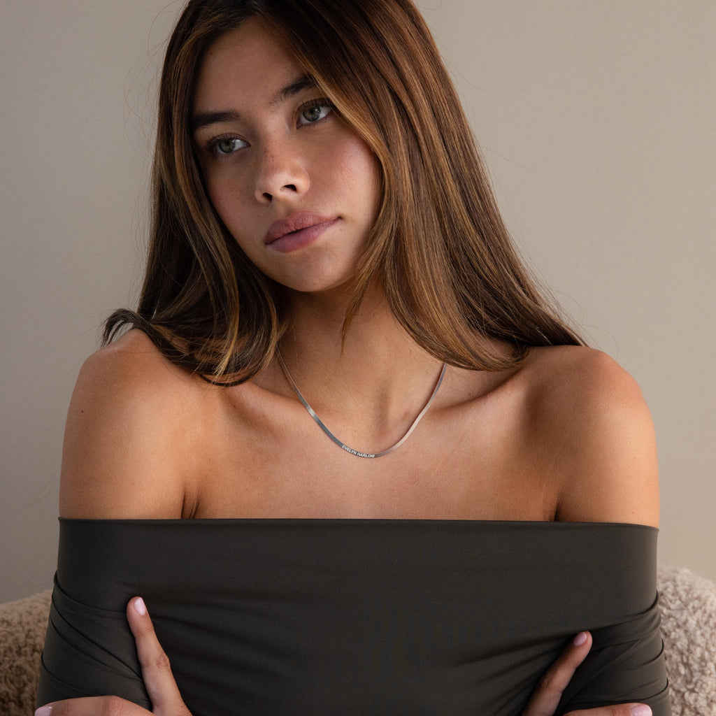 A woman with long brown hair, wearing an off-shoulder dark top and an Engraved Herringbone Name Necklace, looks thoughtful.