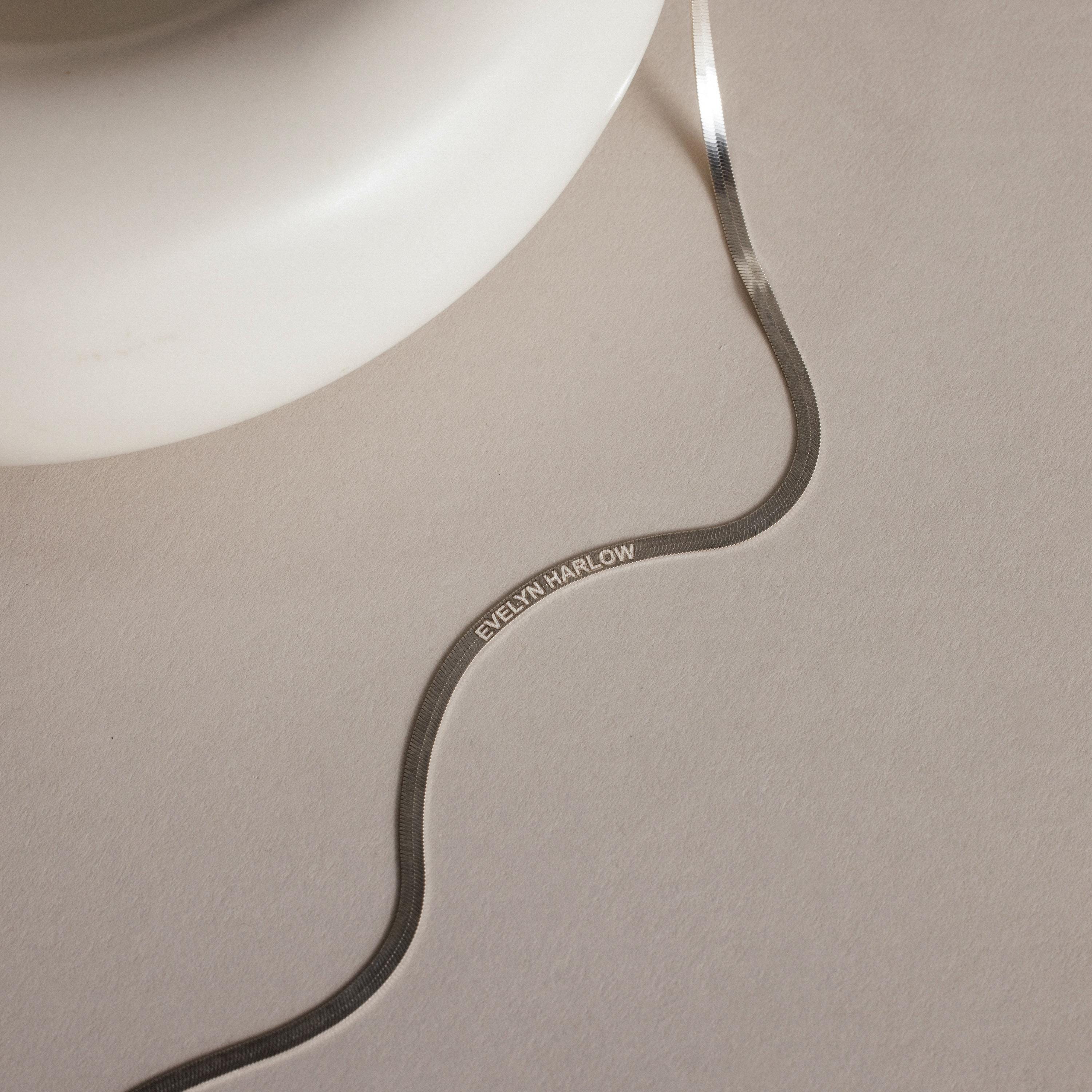 An Engraved Herringbone Name Necklace reading EVELYN HARLOW is displayed on a light beige surface beside a white object.