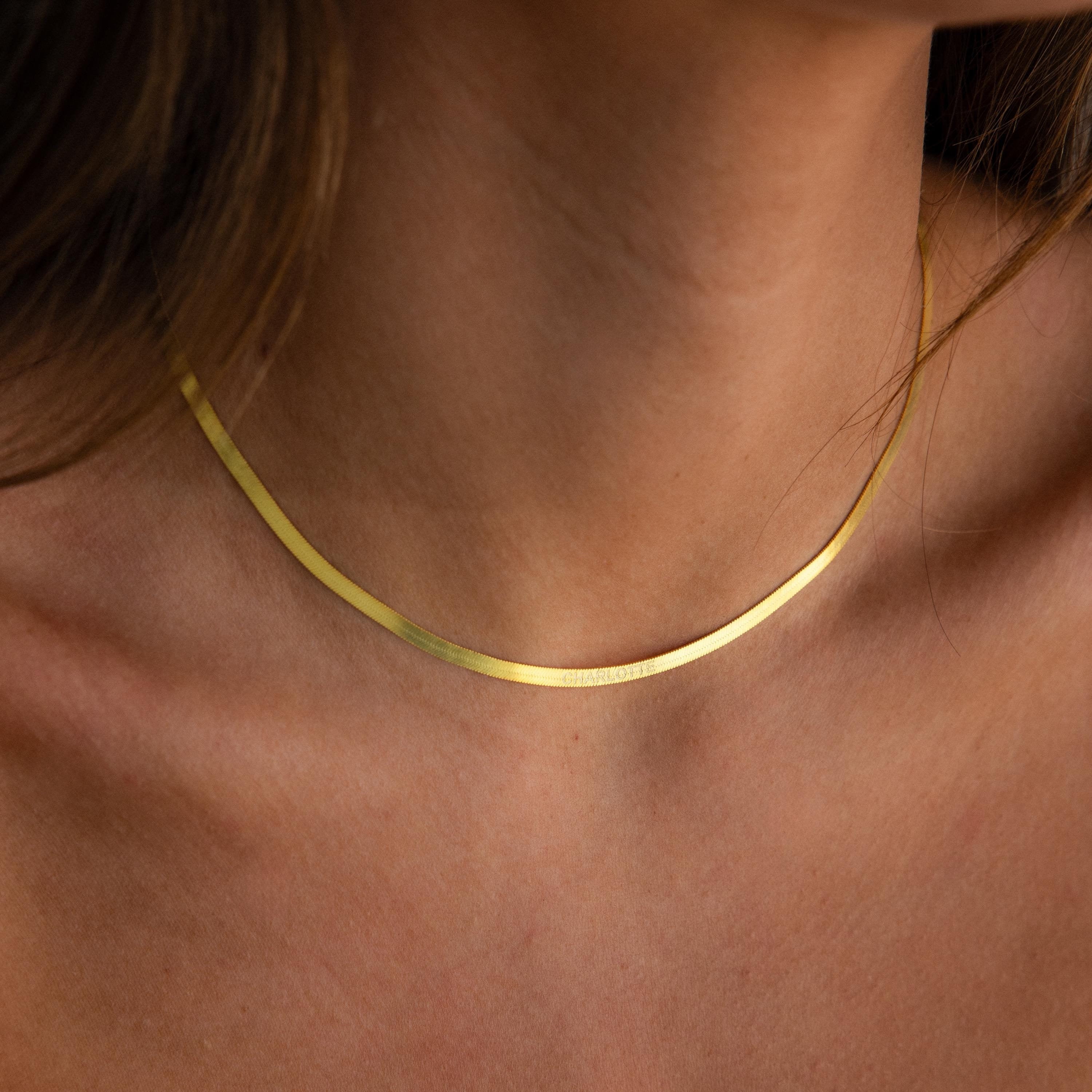 A close-up of a person wearing the Engraved Herringbone Name Necklace, a thin gold chain, on their bare neck and collarbone.