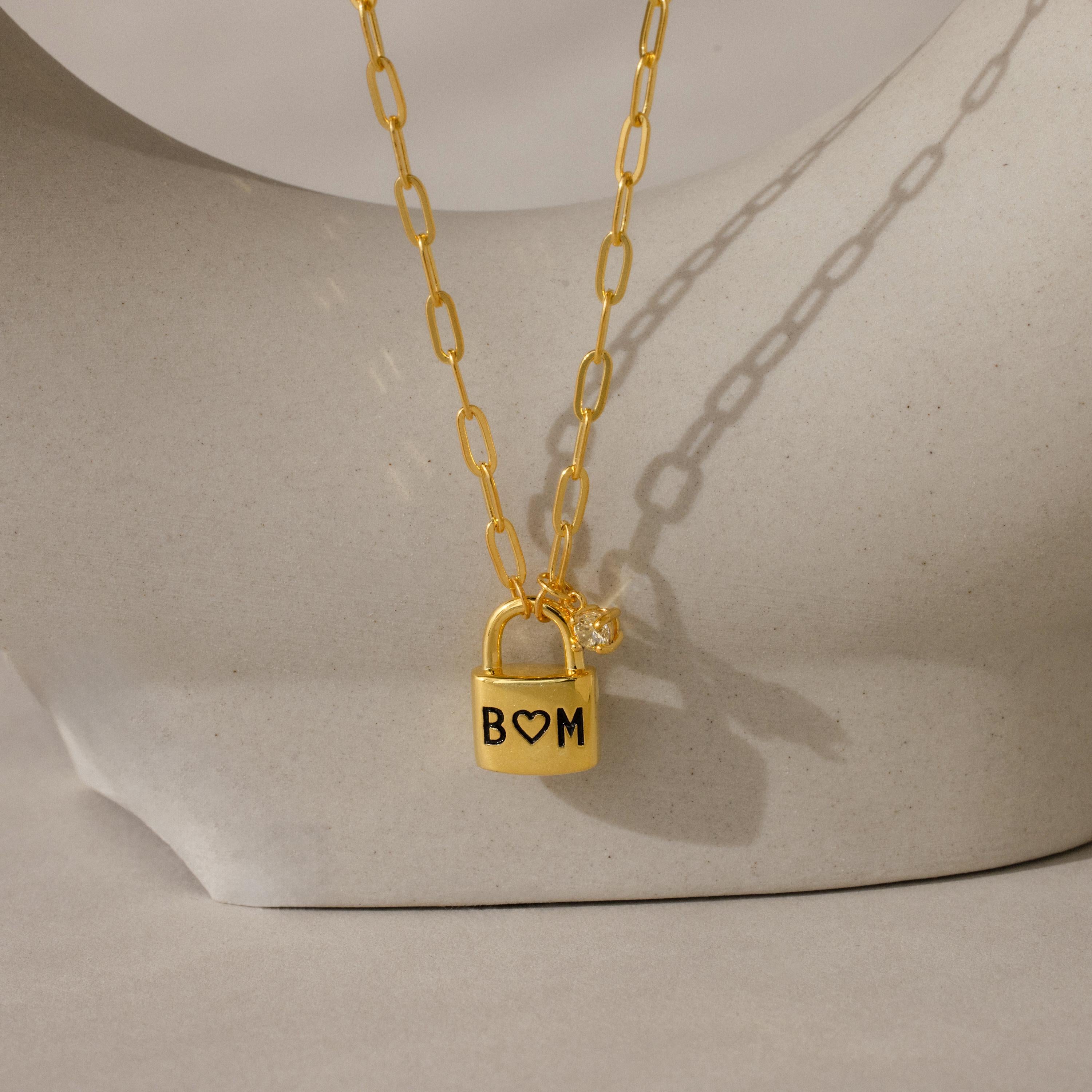 The Initial Birthstone Padlock Necklace features a personalized engraved padlock and a sparkling birthstone charm, elegantly displayed on a sculpture.