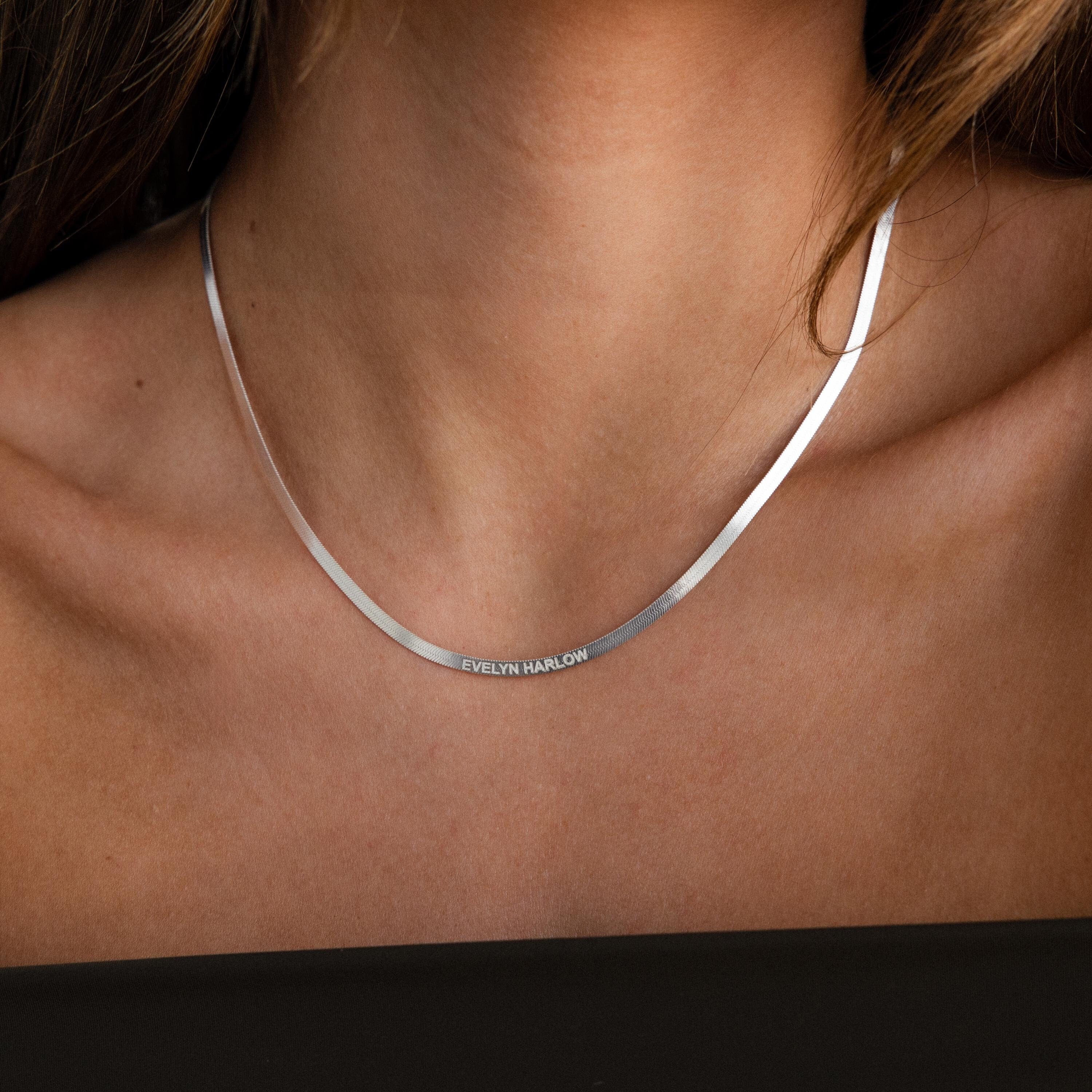 A close-up of a woman wearing the Engraved Herringbone Name Necklace in silver, personalized with EVELYN HARLOW elegantly displayed along its delicate chain.