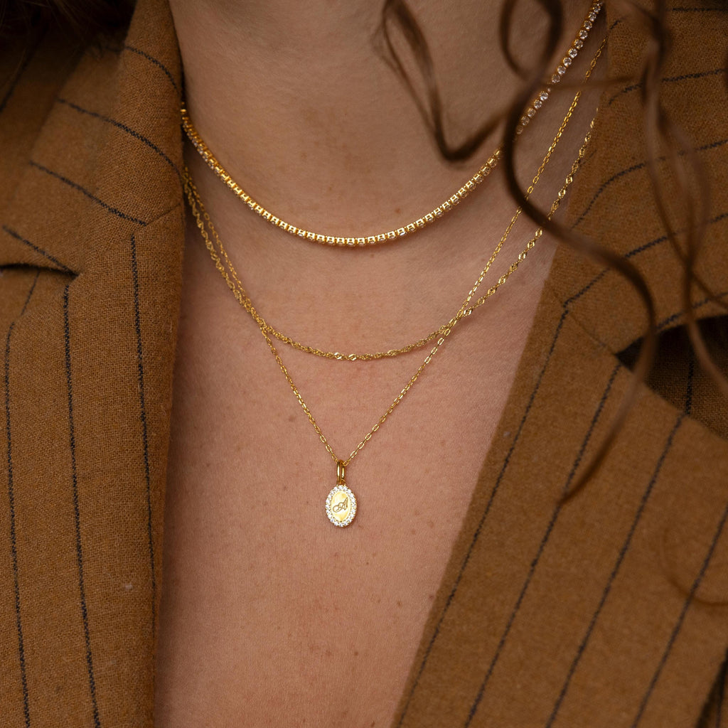 Close-up of layered gold necklaces, featuring the Pave Initial Pendant Necklace, worn by someone in a brown pinstripe blazer with curly hair.