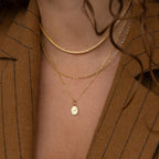Close-up of layered gold necklaces, featuring the Pave Initial Pendant Necklace, worn by someone in a brown pinstripe blazer with curly hair.