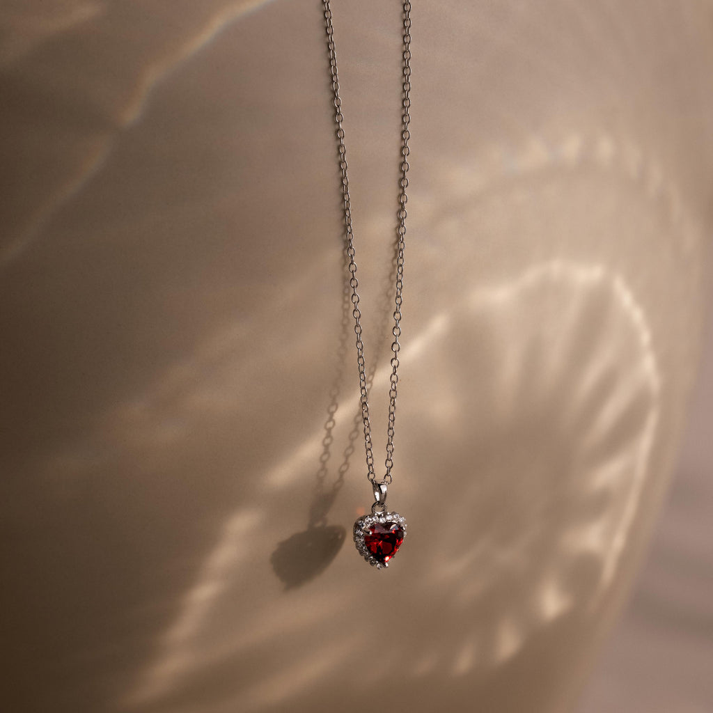 The Pave Garnet Heart Necklace showcases a heart-shaped red gemstone pendant, casting radiant patterns on a beige surface.