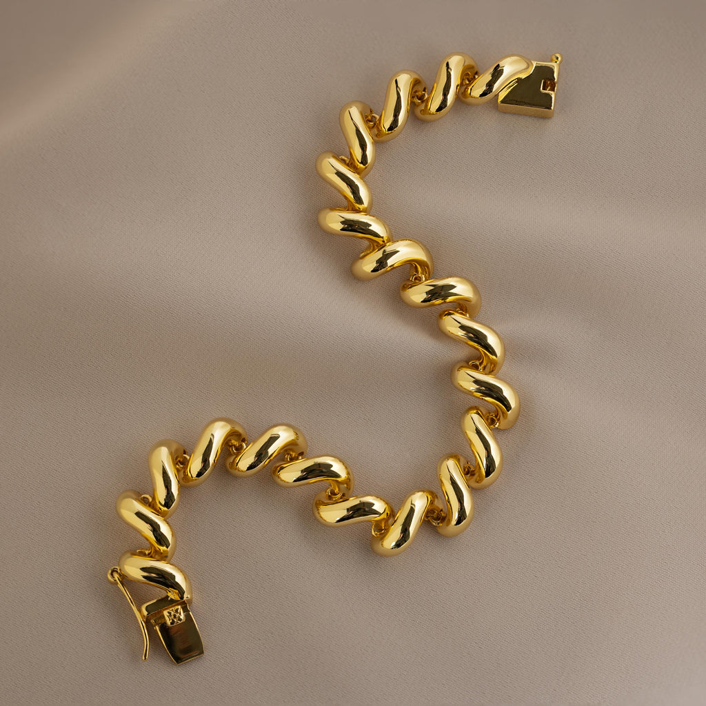 The Twisted Chain Bracelet features bold shining links in a chunky curved S-shaped design, exuding effortless glamour against a beige fabric background.