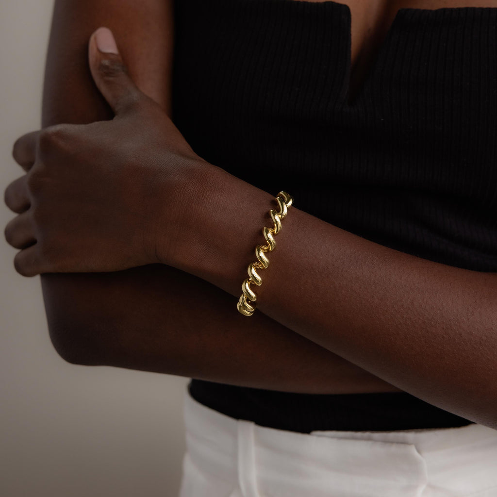 A person wearing the Twisted Chain Bracelet in 18K Gold, paired with a black top and white pants, stands with arms crossed, radiating effortless glamour.