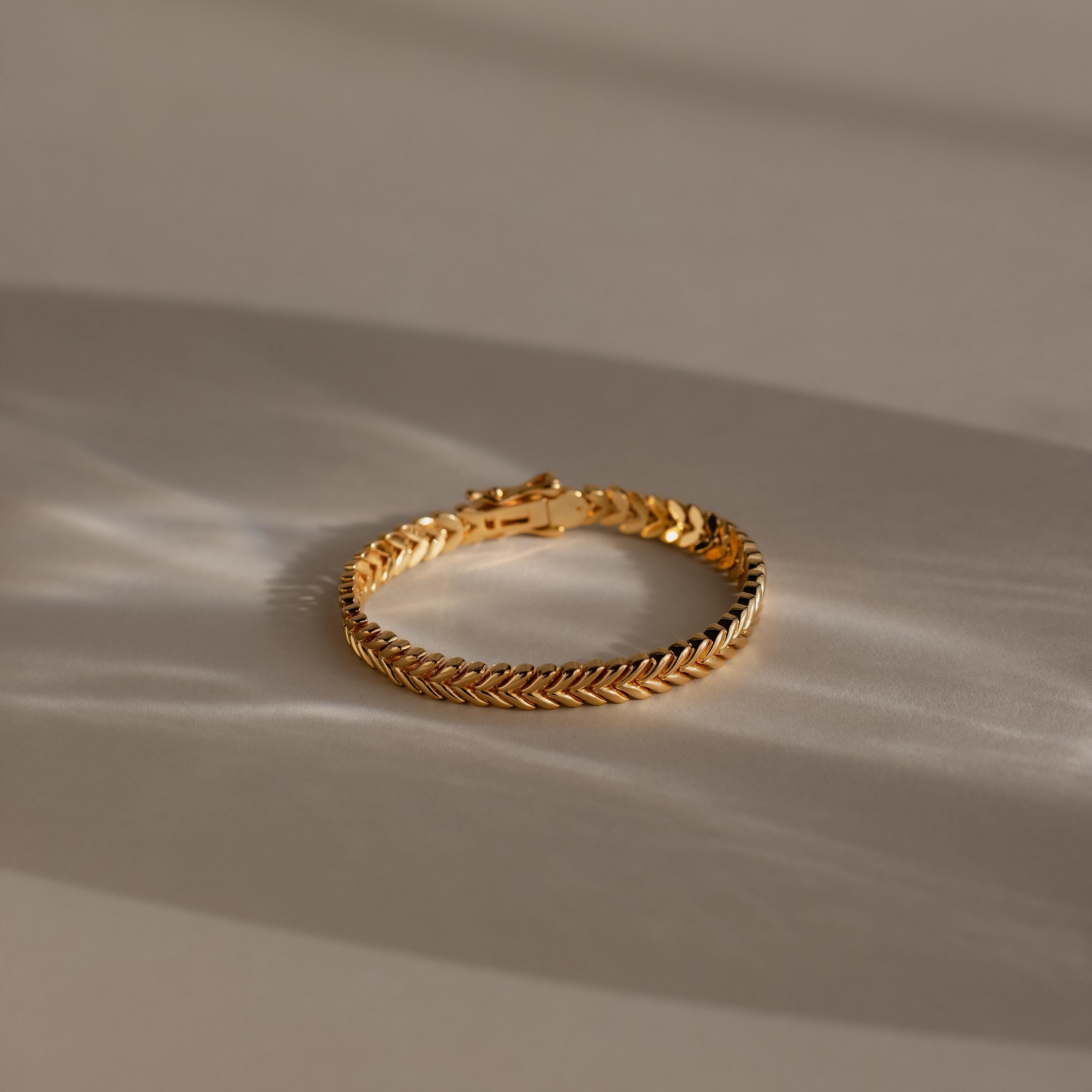 The V-Link Chain Tennis Bracelet, featuring gold sculptural links in a herringbone design, is displayed on a beige surface under soft natural light with gentle shadows.