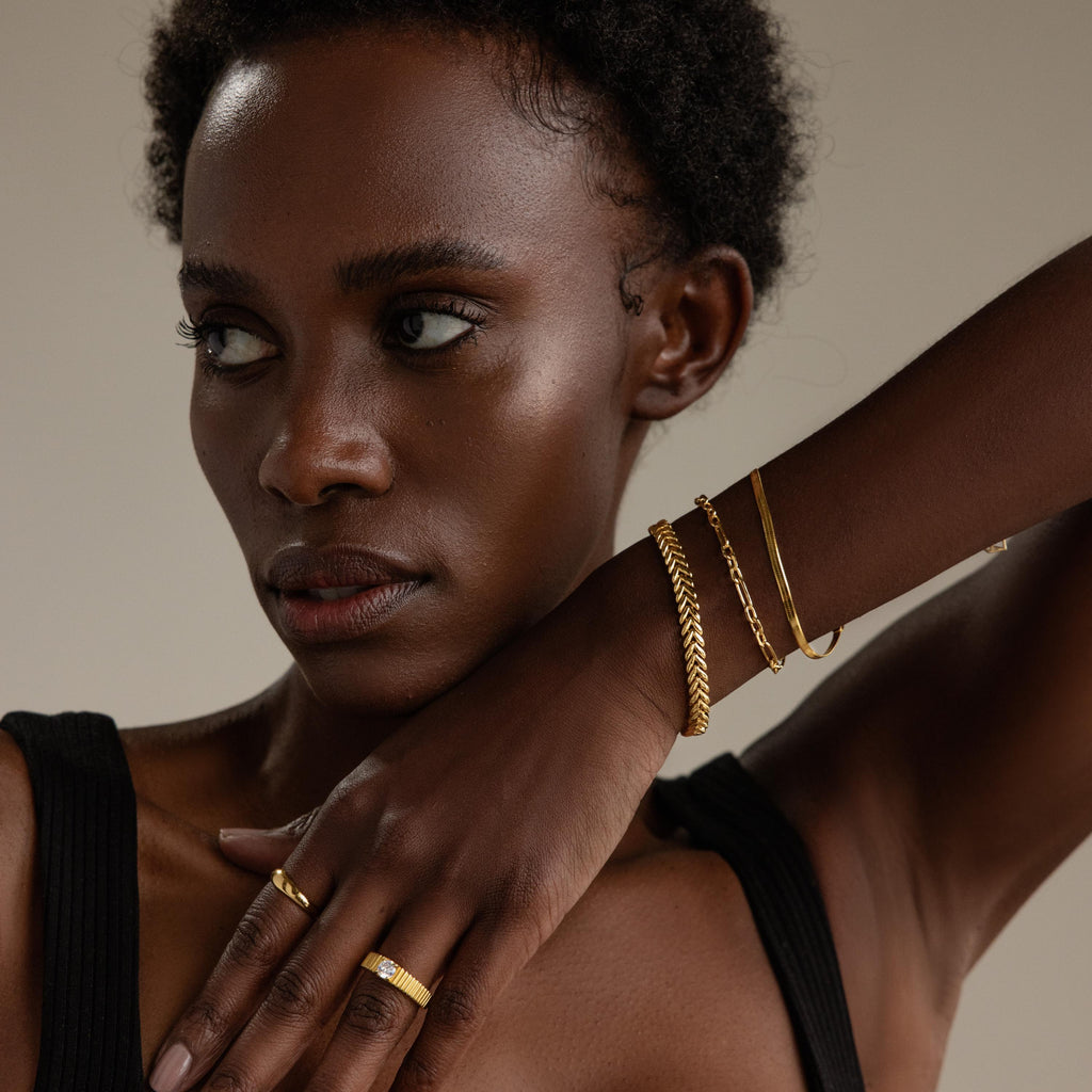 A woman wearing the V-Link Chain Tennis Bracelet and sculptural gold jewelry rests her hand on her shoulder while looking away.