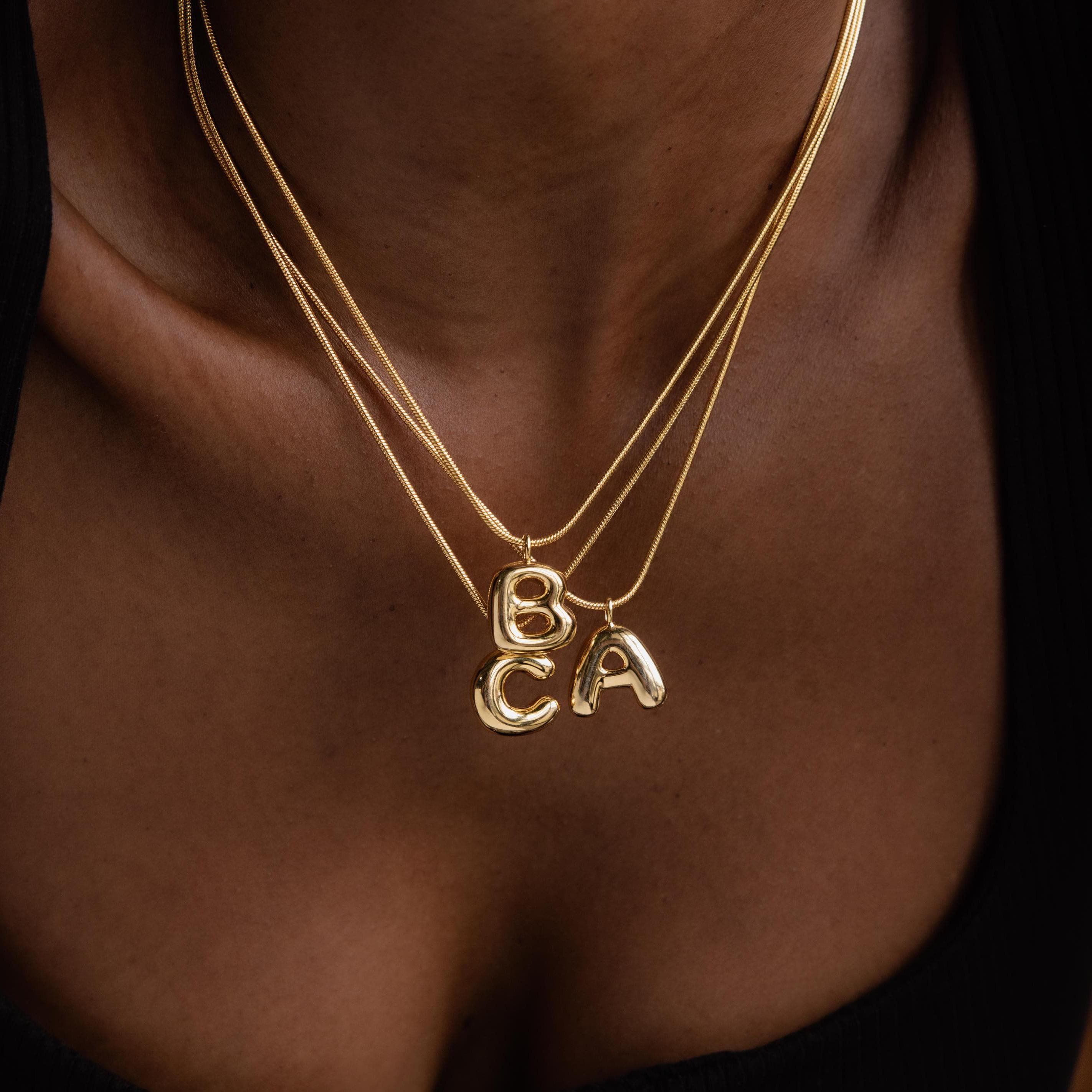 Three Bubble Letter Necklaces in 18K Gold, featuring chunky letter charm pendants B, C, and A, rest against dark clothing—ideal for anyone who loves personalized jewelry.