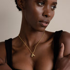 A woman wearing the Bubble Letter Necklace in 18K Gold with an “A” pendant looks to the side against a neutral background, showcasing her personalized jewelry.