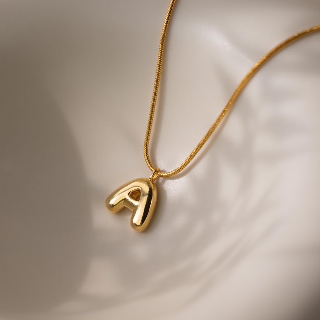 Bubble Letter Necklace in 18K Gold featuring a shiny, chunky letter A pendant displayed on a smooth, light-colored surface.