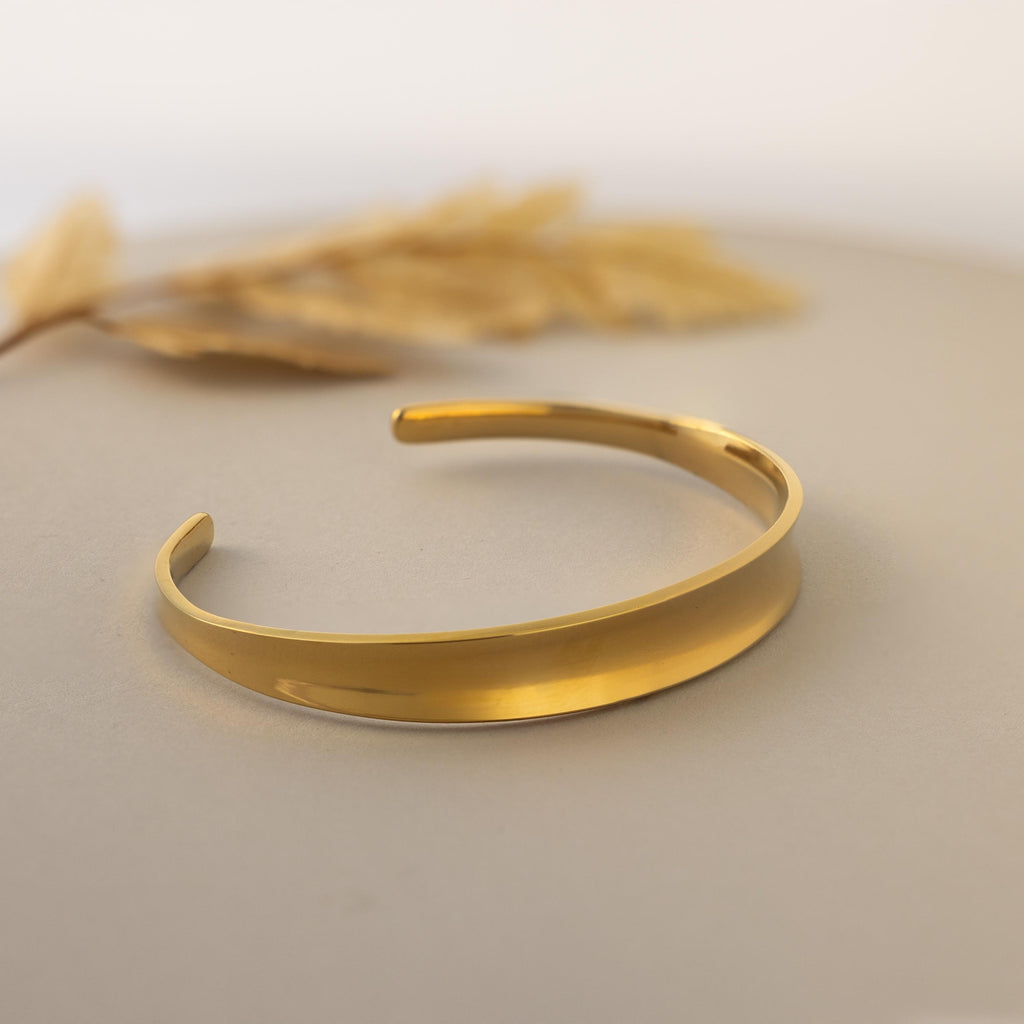 The Dome Cuff Bracelet is displayed on a neutral surface with dried grass in the background, showcasing its simple gold design that adds a stylish accent to any look.