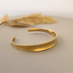 The Dome Cuff Bracelet is displayed on a neutral surface with dried grass in the background, showcasing its simple gold design that adds a stylish accent to any look.