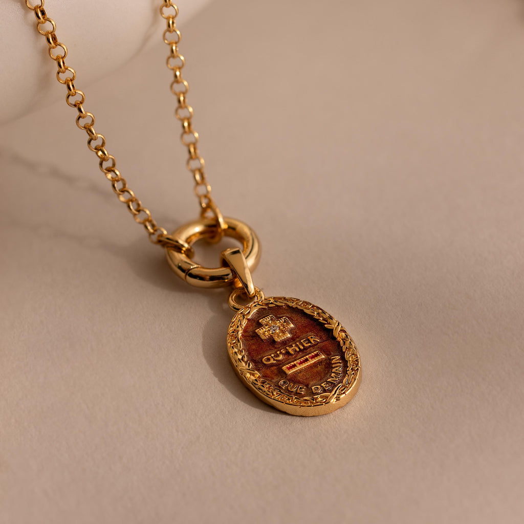 The Love Poem Pendant Necklace features an oval pendant engraved with a crest, presented on a gold chain—a timeless symbol of love, shown resting on a beige surface.
