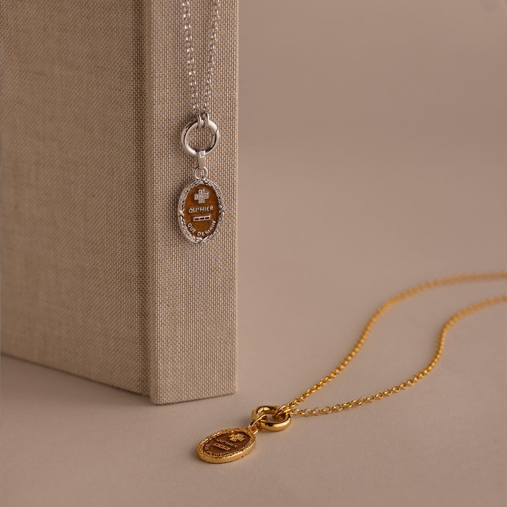 The Love Poem Pendant Necklace, available in silver and gold with elegant oval pendants, is shown resting on and next to a beige textured book—a timeless symbol of love.
