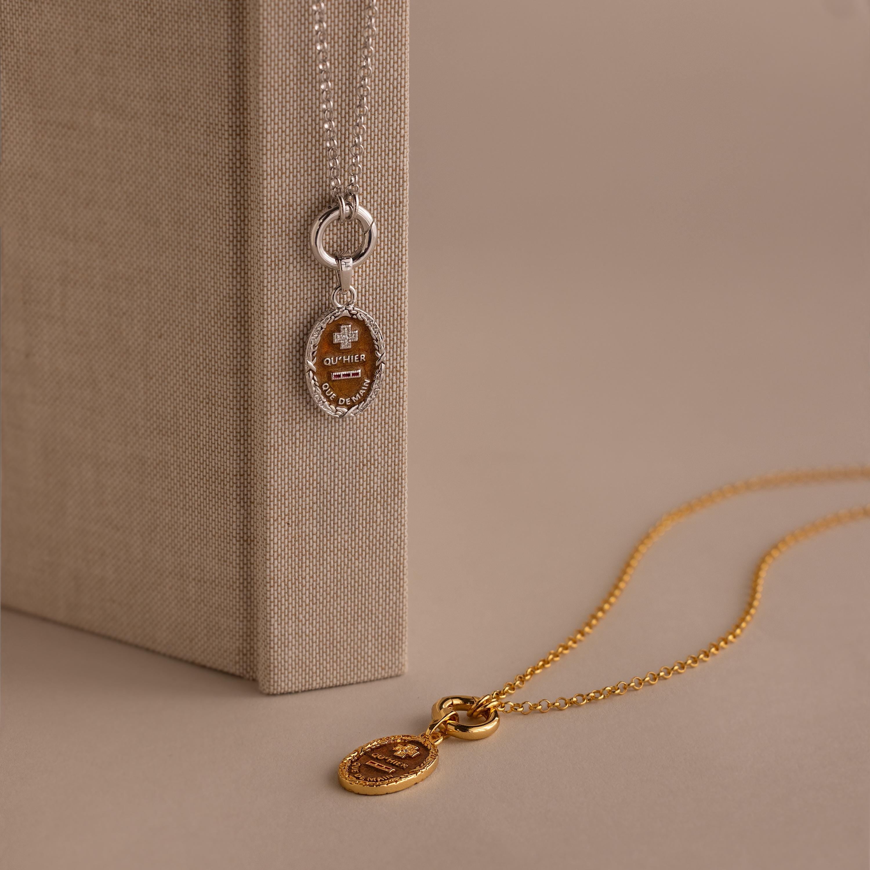 The Love Poem Pendant Necklace, available in silver and gold with elegant oval pendants, is shown resting on and next to a beige textured book—a timeless symbol of love.
