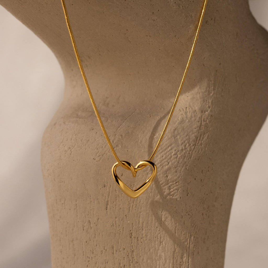 The Heart Knot Necklace features a gold open heart charm on a sleek snake chain, elegantly displayed on a textured beige surface under soft lighting.
