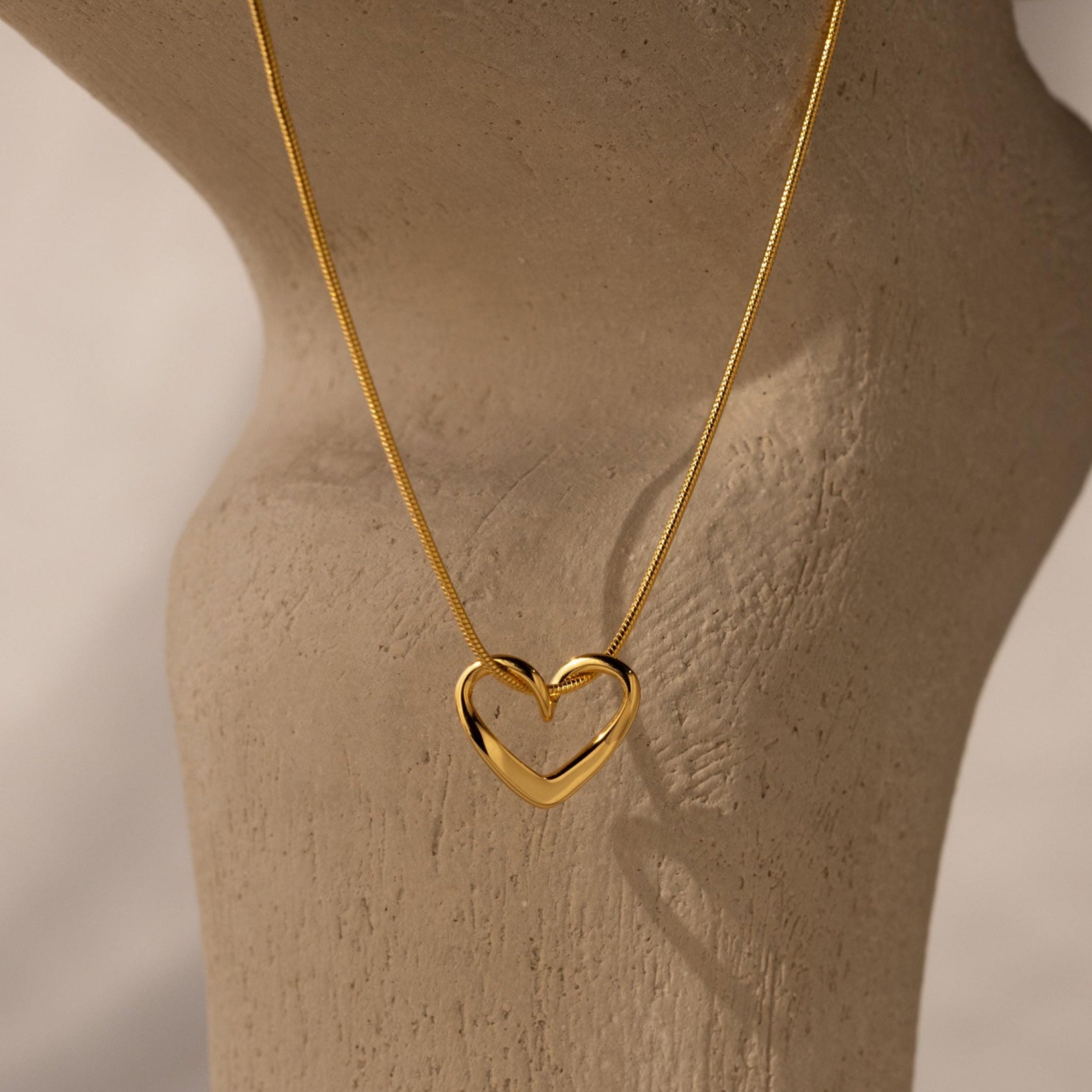 The Heart Knot Necklace features a gold open heart charm on a sleek snake chain, elegantly displayed on a textured beige surface under soft lighting.