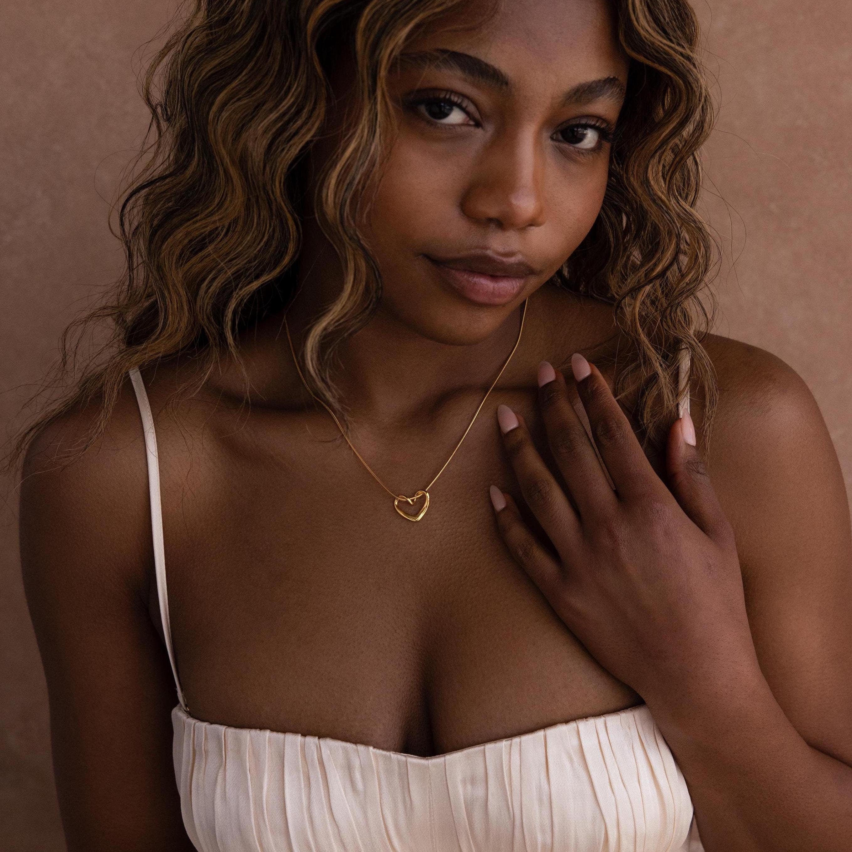 A woman with wavy hair in a cream dress wears the Heart Knot Necklace—a gold open heart charm on a delicate snake chain—and rests her hand on her chest.