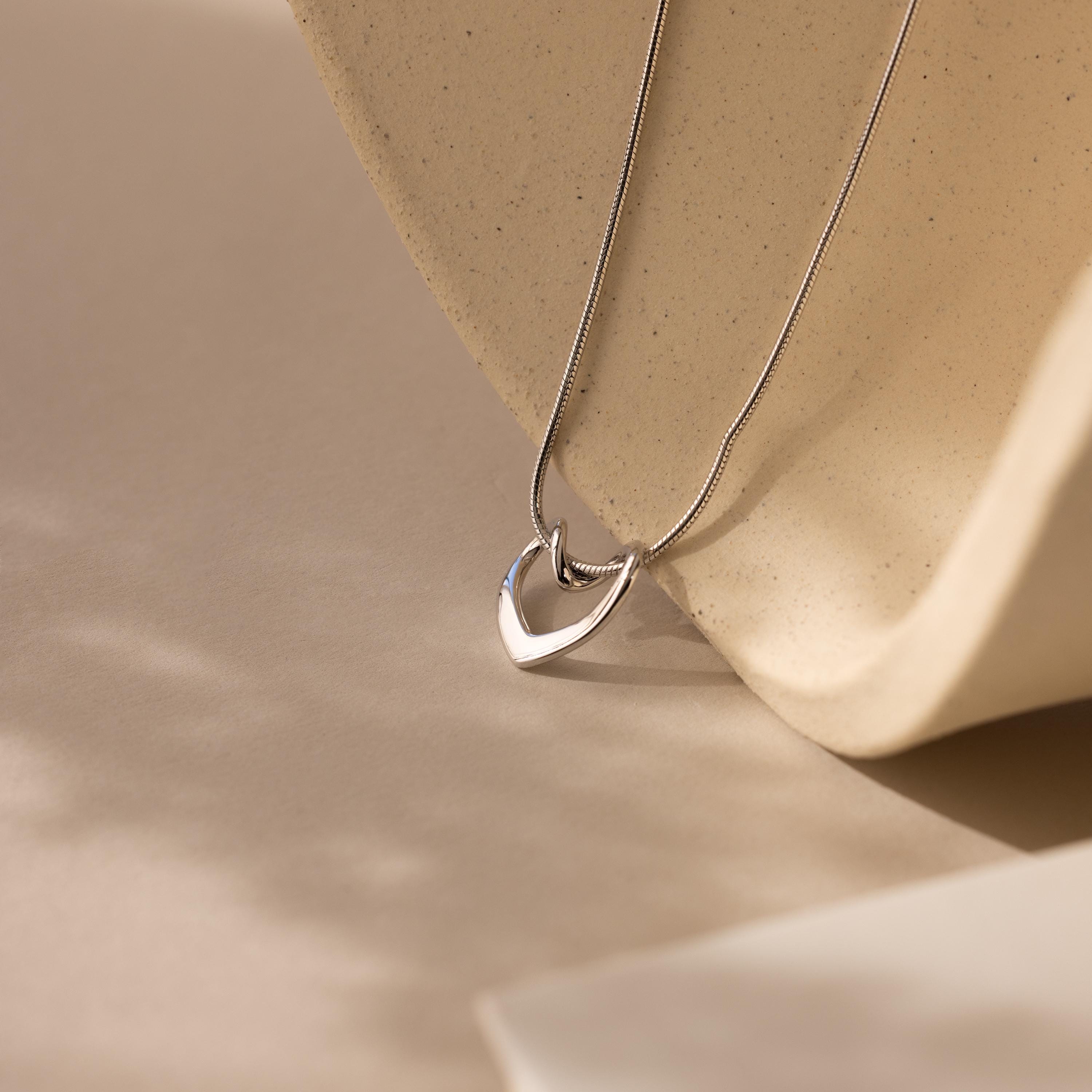 The Heart Knot Necklace features a silver heart-shaped pendant on a delicate snake chain, presented against a beige textured background with soft natural light and gentle shadows.