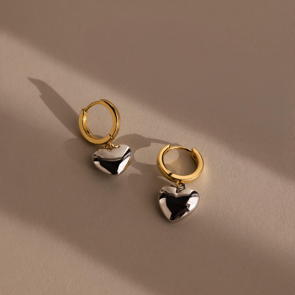 The Mixed Metal Heart Huggies feature gold hoops with silver puff heart charms, displayed on a beige surface with soft shadows to highlight their elegant, contemporary style.