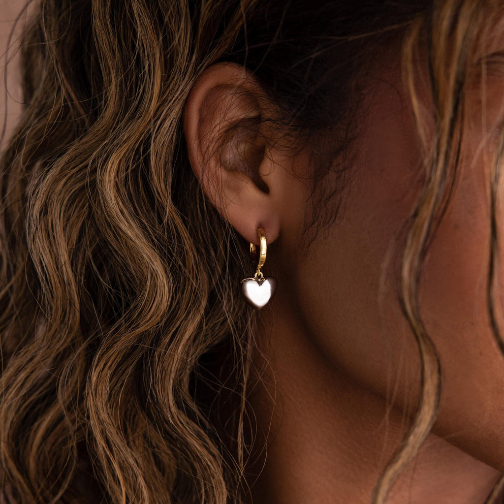 Close-up of a person with wavy hair wearing Mixed Metal Heart Huggies—gold hoop earrings with heart-shaped charms.