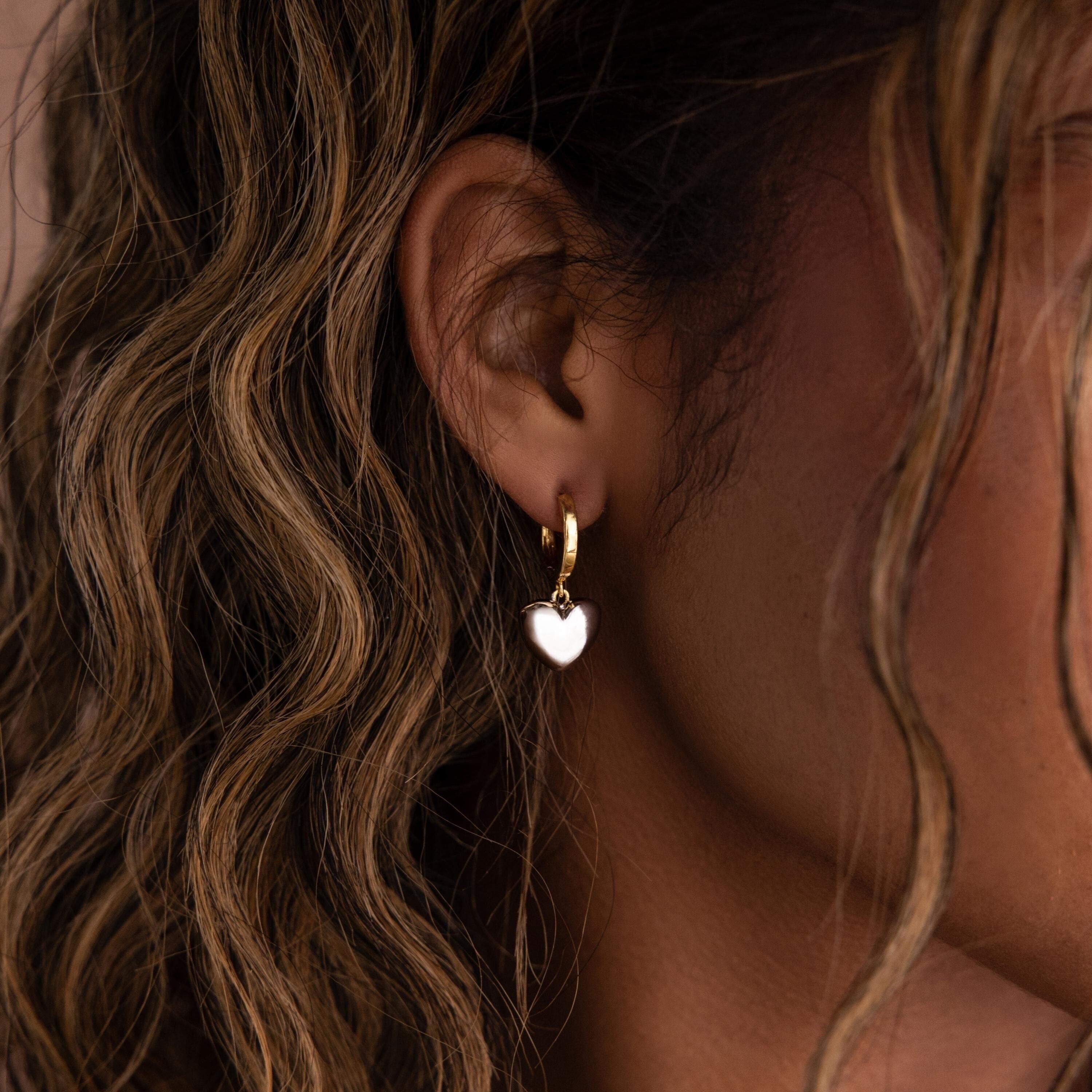 Close-up of a person with wavy hair wearing Mixed Metal Heart Huggies—gold hoop earrings with heart-shaped charms.