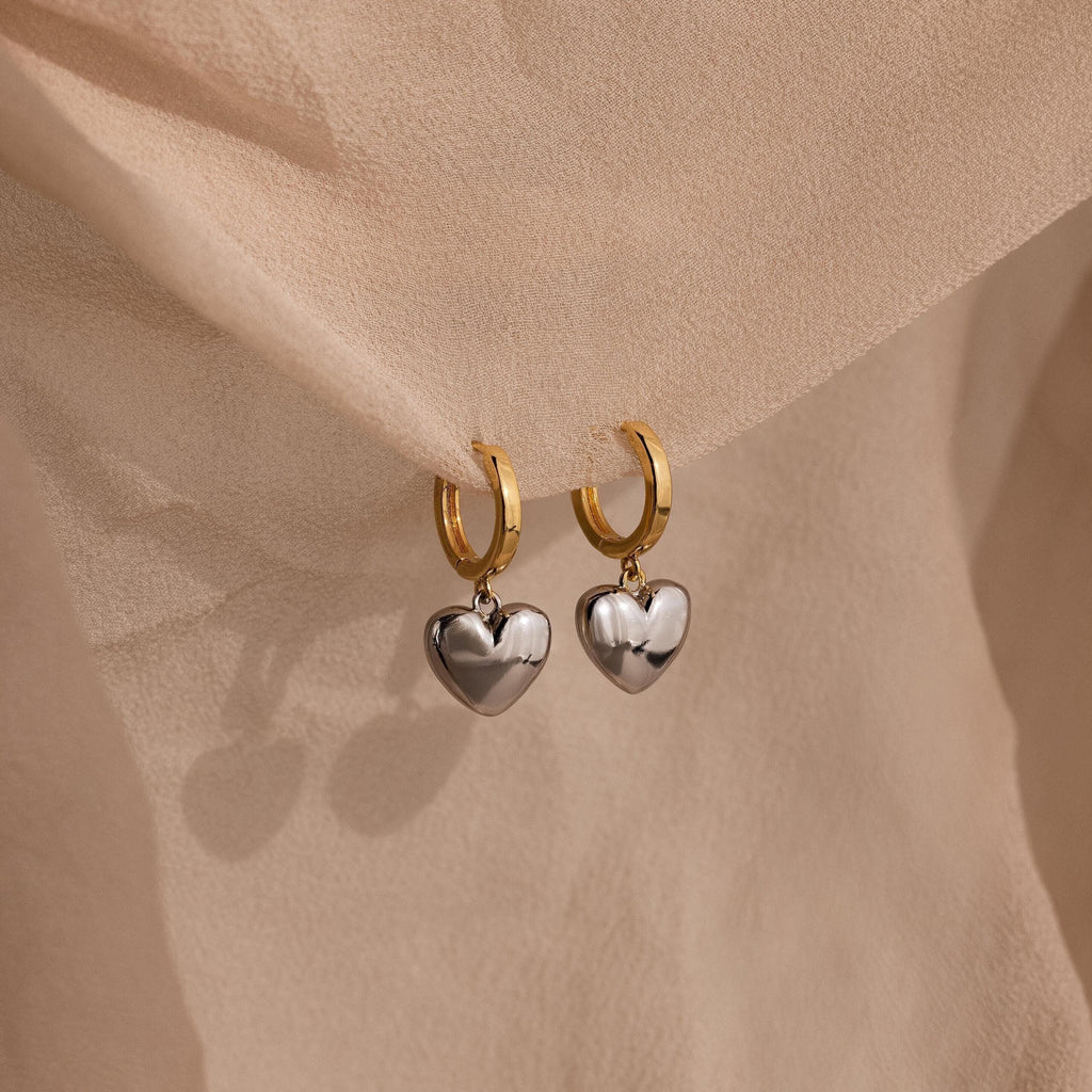 The Mixed Metal Heart Huggies feature gold hoops with silver puff heart charms, displayed on soft beige fabric for a stylish mixed metal look.