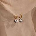 The Mixed Metal Heart Huggies feature gold hoops with silver puff heart charms, displayed on soft beige fabric for a stylish mixed metal look.