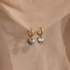 The Mixed Metal Heart Huggies feature gold hoops with silver puff heart charms, displayed on soft beige fabric for a stylish mixed metal look.