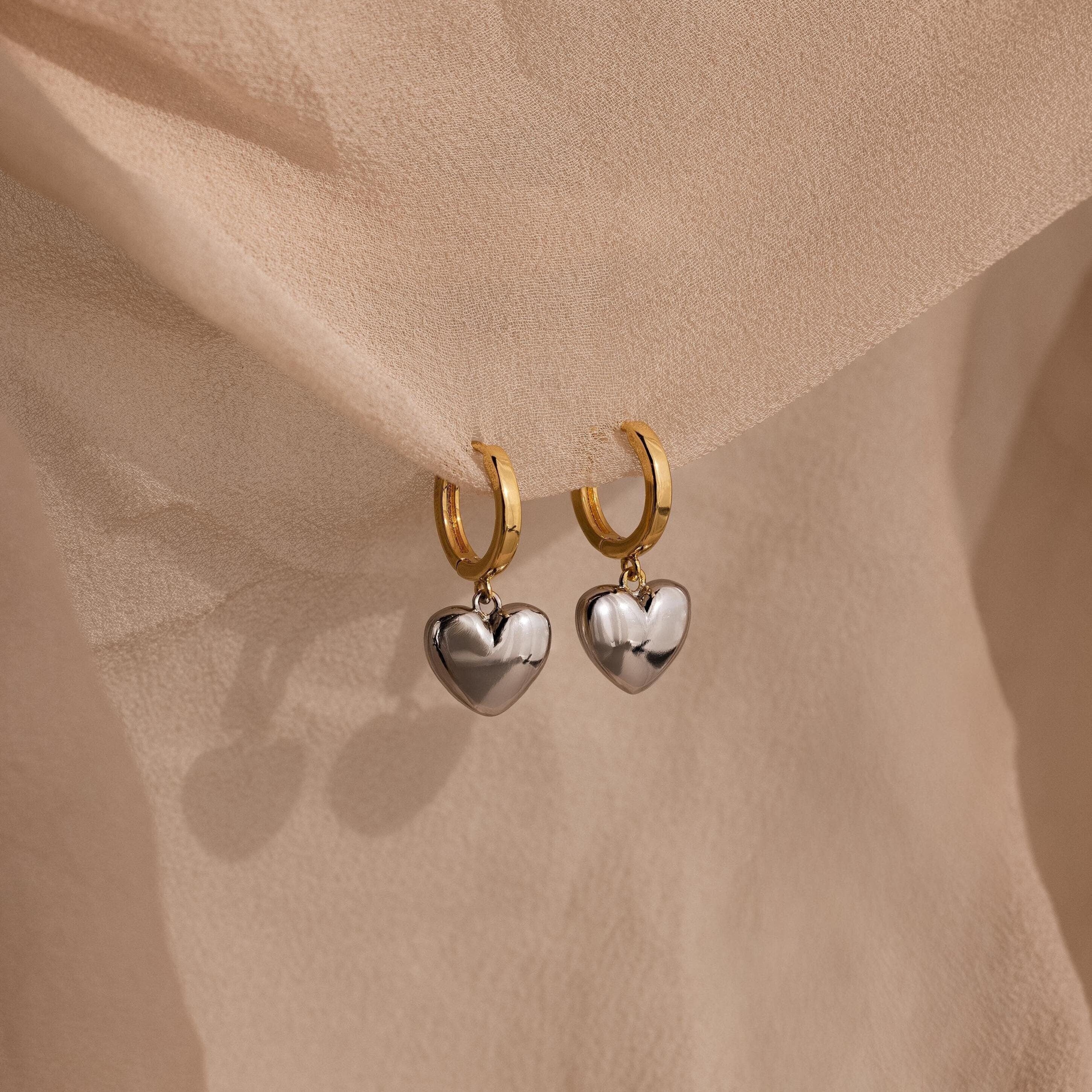 The Mixed Metal Heart Huggies feature gold hoops with silver puff heart charms, displayed on soft beige fabric for a stylish mixed metal look.