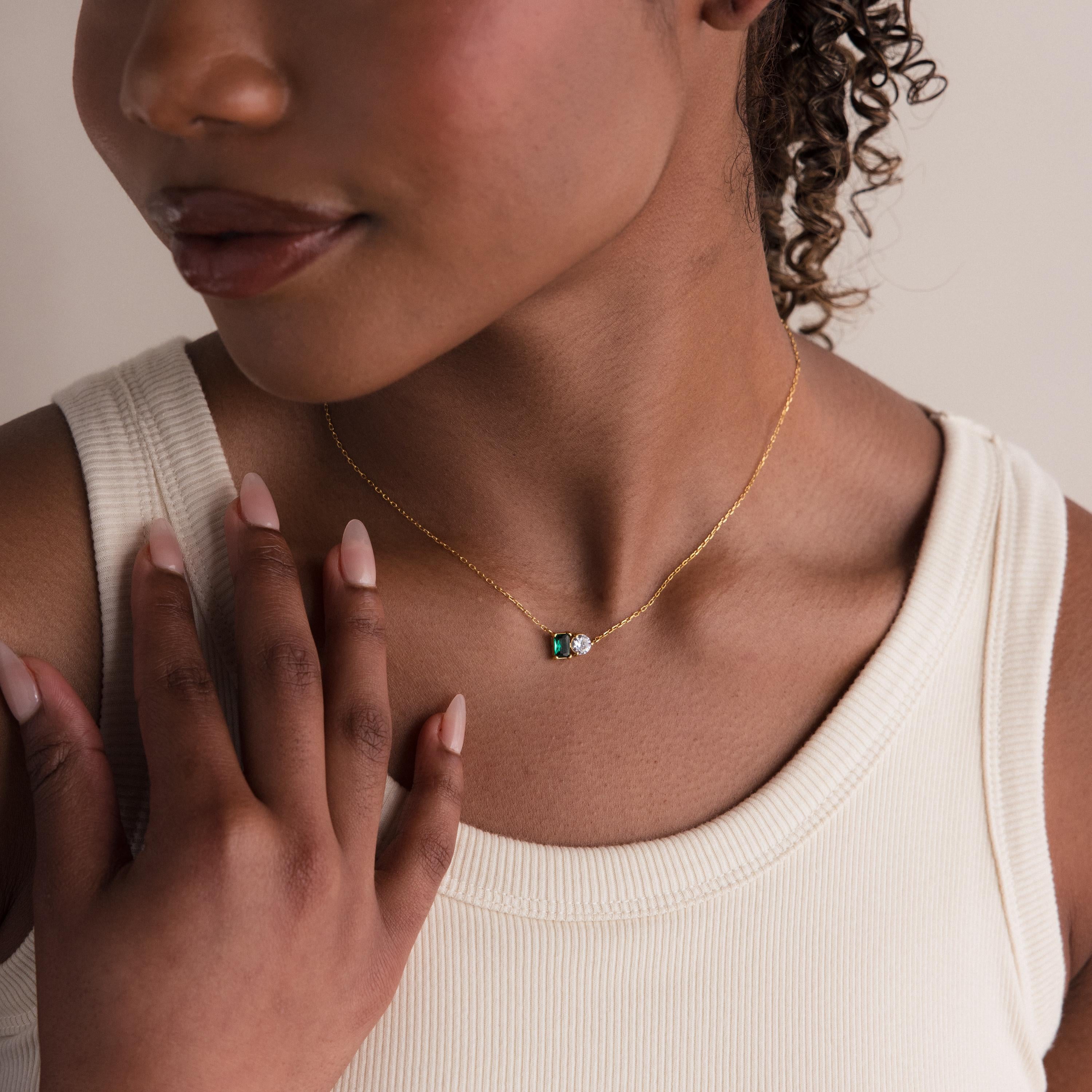 A woman in a cream tank top wears the Toi et Moi Emerald Birthstone Necklace, featuring a delicate gold chain with a small green and white pendant.