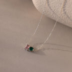 The Toi et Moi Emerald Birthstone Necklace features pink and green gemstones, making it a perfect personalized gift. Displayed on a soft beige surface.