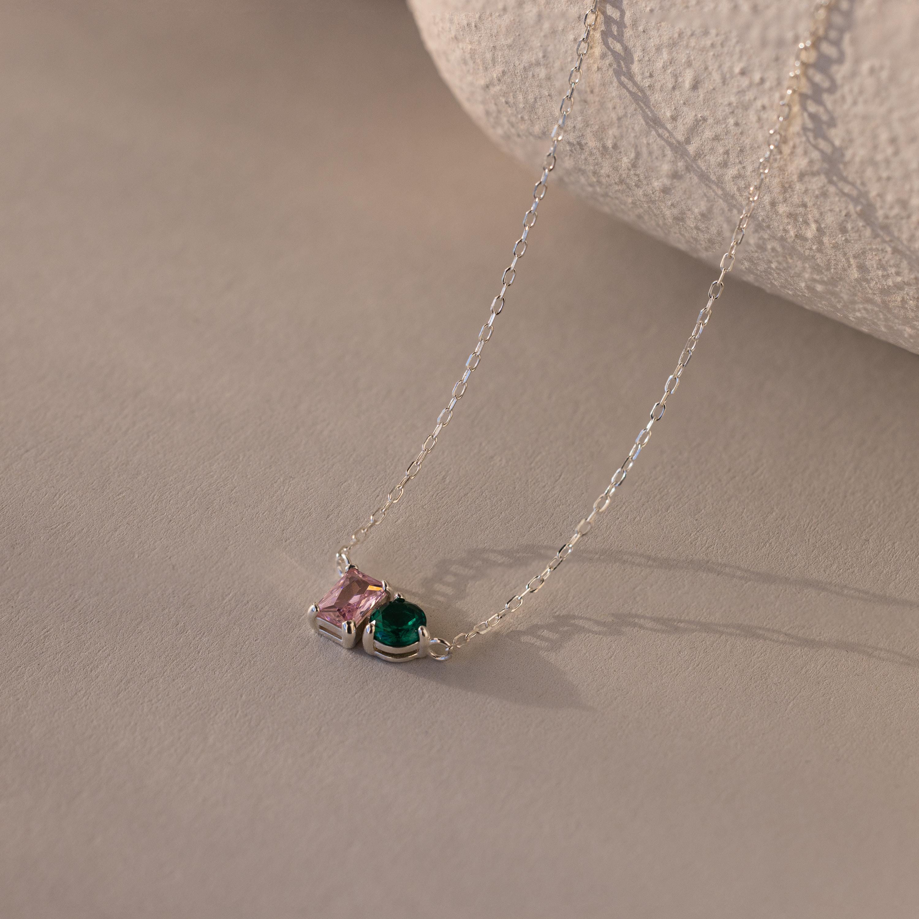 The Toi et Moi Emerald Birthstone Necklace features pink and green gemstones, making it a perfect personalized gift. Displayed on a soft beige surface.
