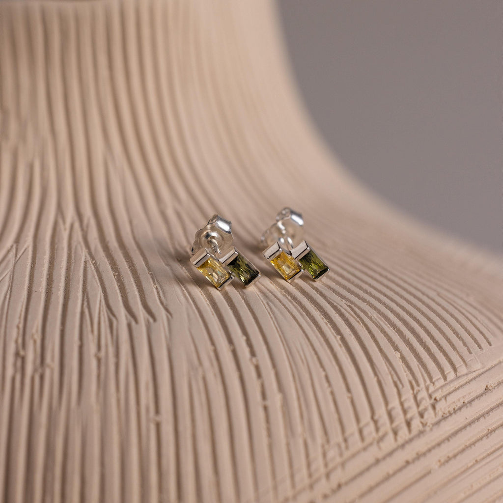 The Duo Baguette Birthstone Studs are elegant silver earrings featuring small green square gemstones, displayed on a textured beige surface—perfect for adding a subtle pop of color to any outfit.