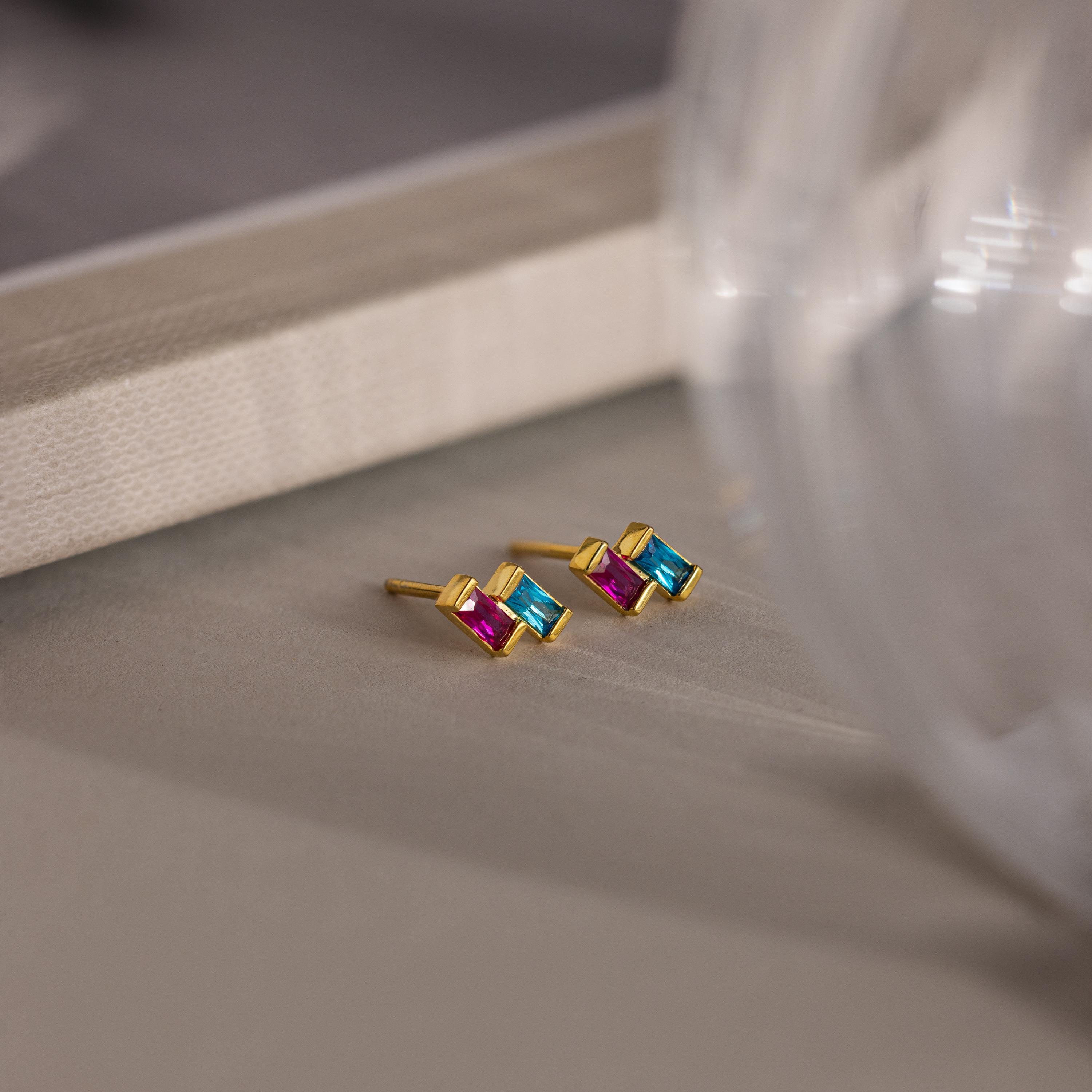The Duo Baguette Birthstone Studs feature gold settings with pink and blue rectangular gemstones, adding a colorful, elegant touch to any outfit.