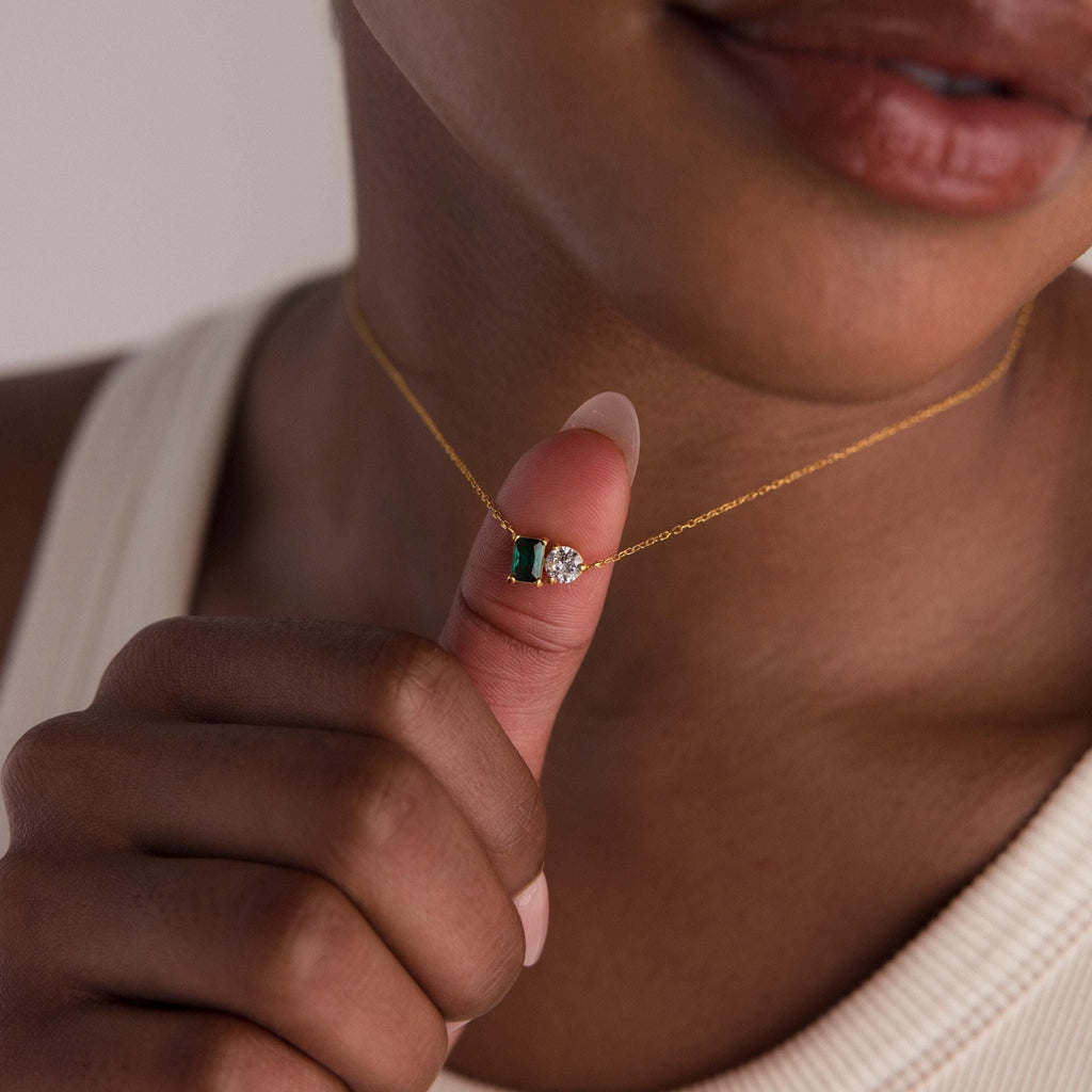 A person holds the Toi et Moi Emerald Birthstone Necklace, adorned with green and clear gemstones, close to their lips.