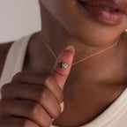 A person holds the Toi et Moi Emerald Birthstone Necklace, adorned with green and clear gemstones, close to their lips.