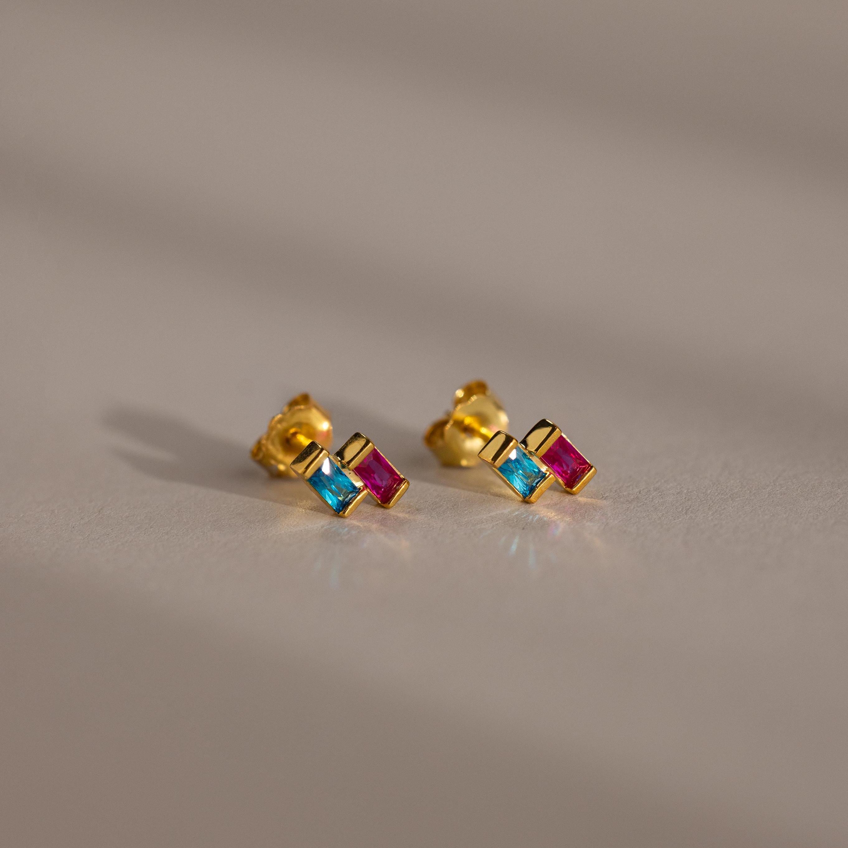 Duo Baguette Birthstone Studs with blue and pink rectangular stones rest on a soft beige surface—elegant, personalized gold earrings perfect for jewelry lovers.