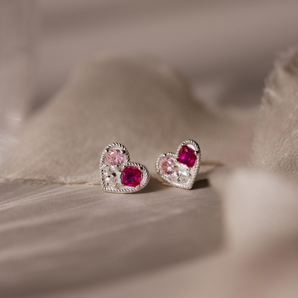 Mixed Birthstone Heart Studs feature silver heart-shaped earrings with customizable pink and red gemstones, beautifully displayed on soft beige fabric.