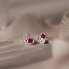 Mixed Birthstone Heart Studs feature silver heart-shaped earrings with customizable pink and red gemstones, beautifully displayed on soft beige fabric.
